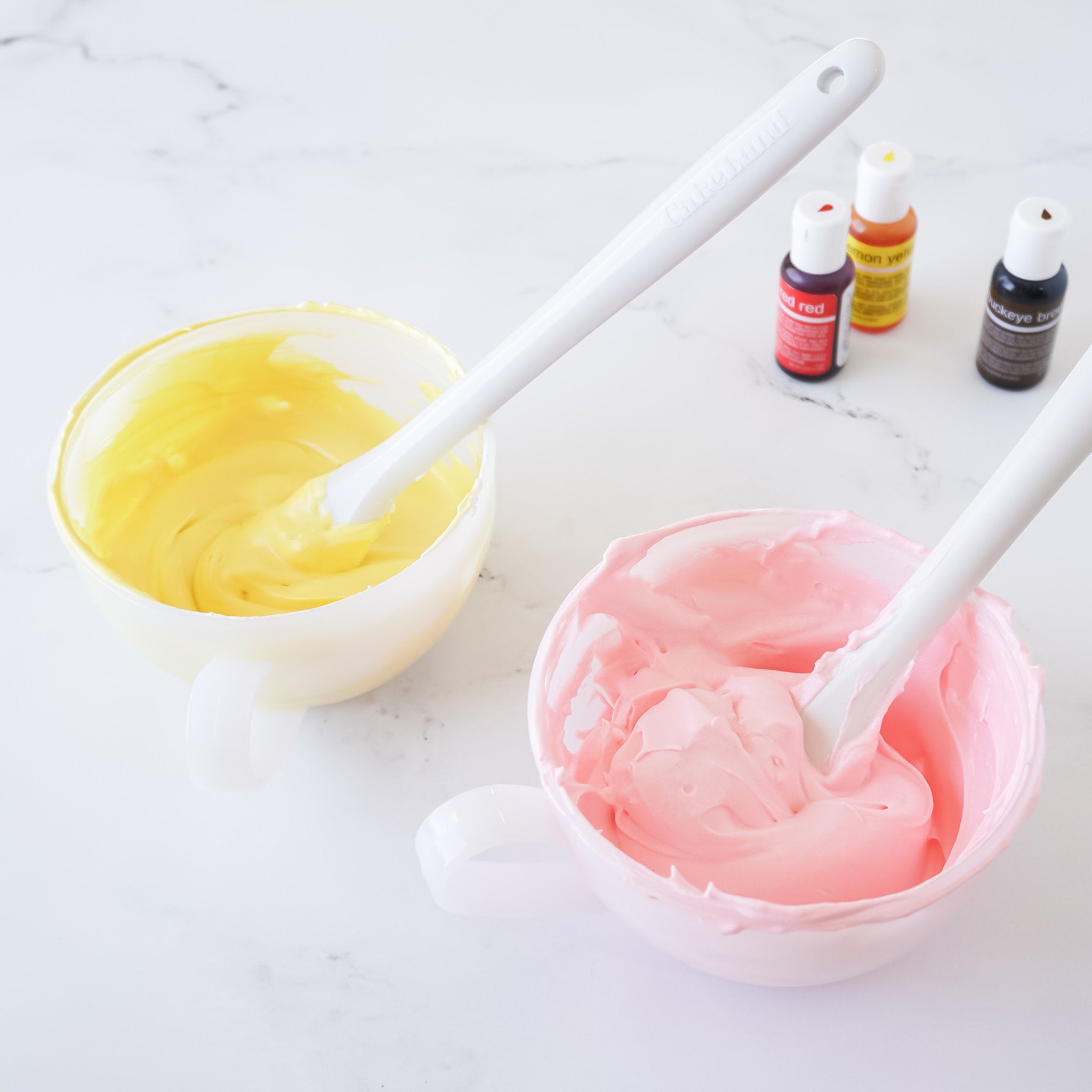 Two white bowls with handles, one filled with yellow frosting and the other filled with pink frosting. A white spatula is in each bowl. Three bottles of food coloring are in the background, one red, one yellow, and one brown.