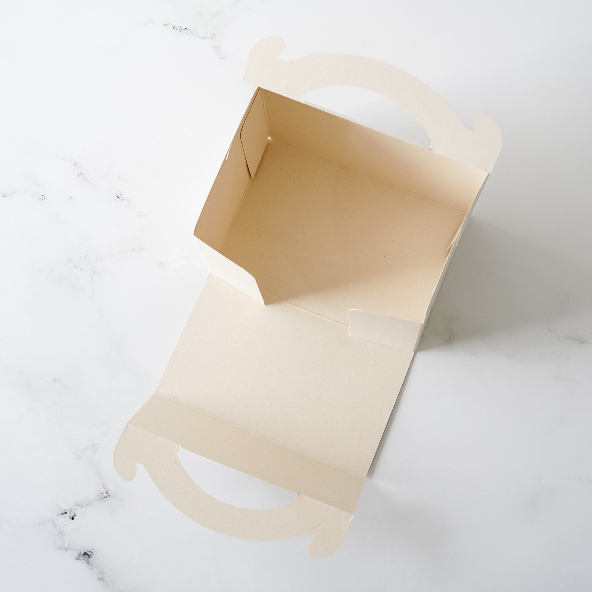 A light beige cardboard box with cut-out handles. The box is open and the inside is empty.