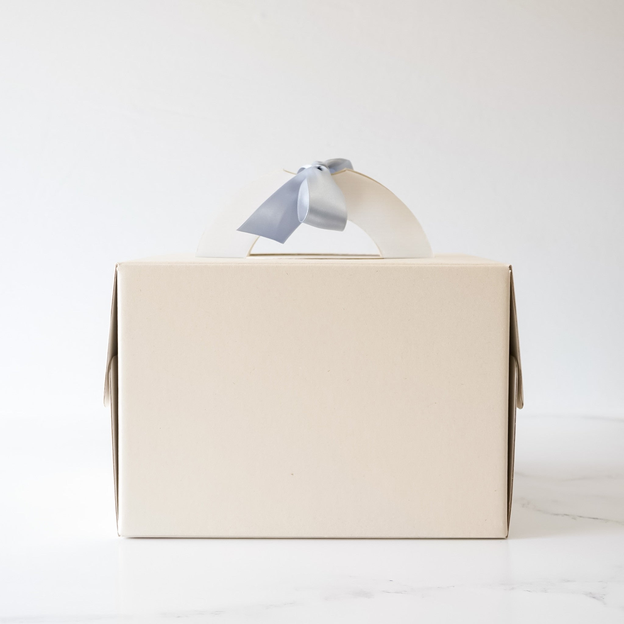 A beige cardboard box with a light blue ribbon tied around the handle. The box is designed for carrying cakes or other baked goods.