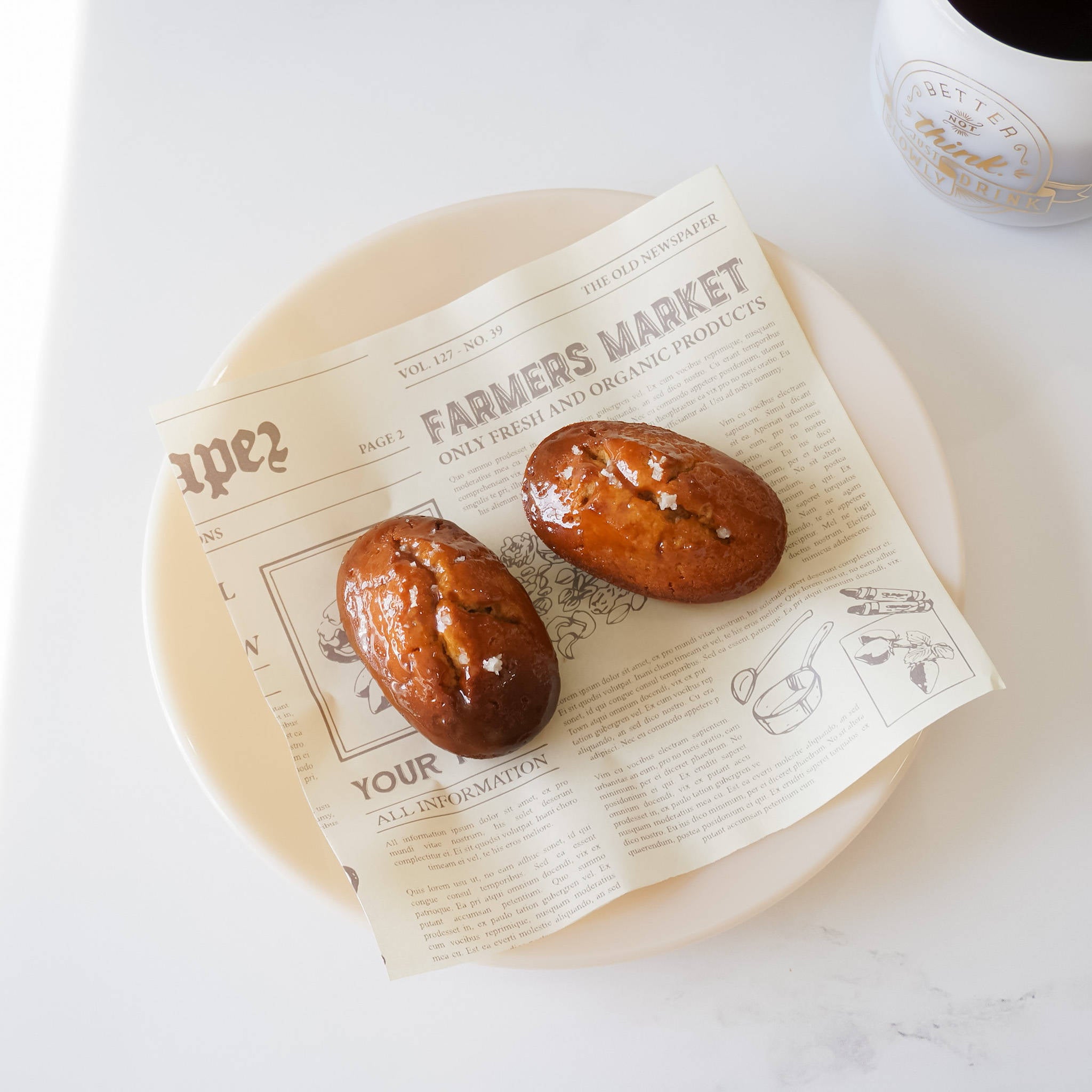 Two small, light brown pastries with a shiny glaze on top. The pastries are on a white plate with a piece of newspaper underneath. The newspaper has the text &