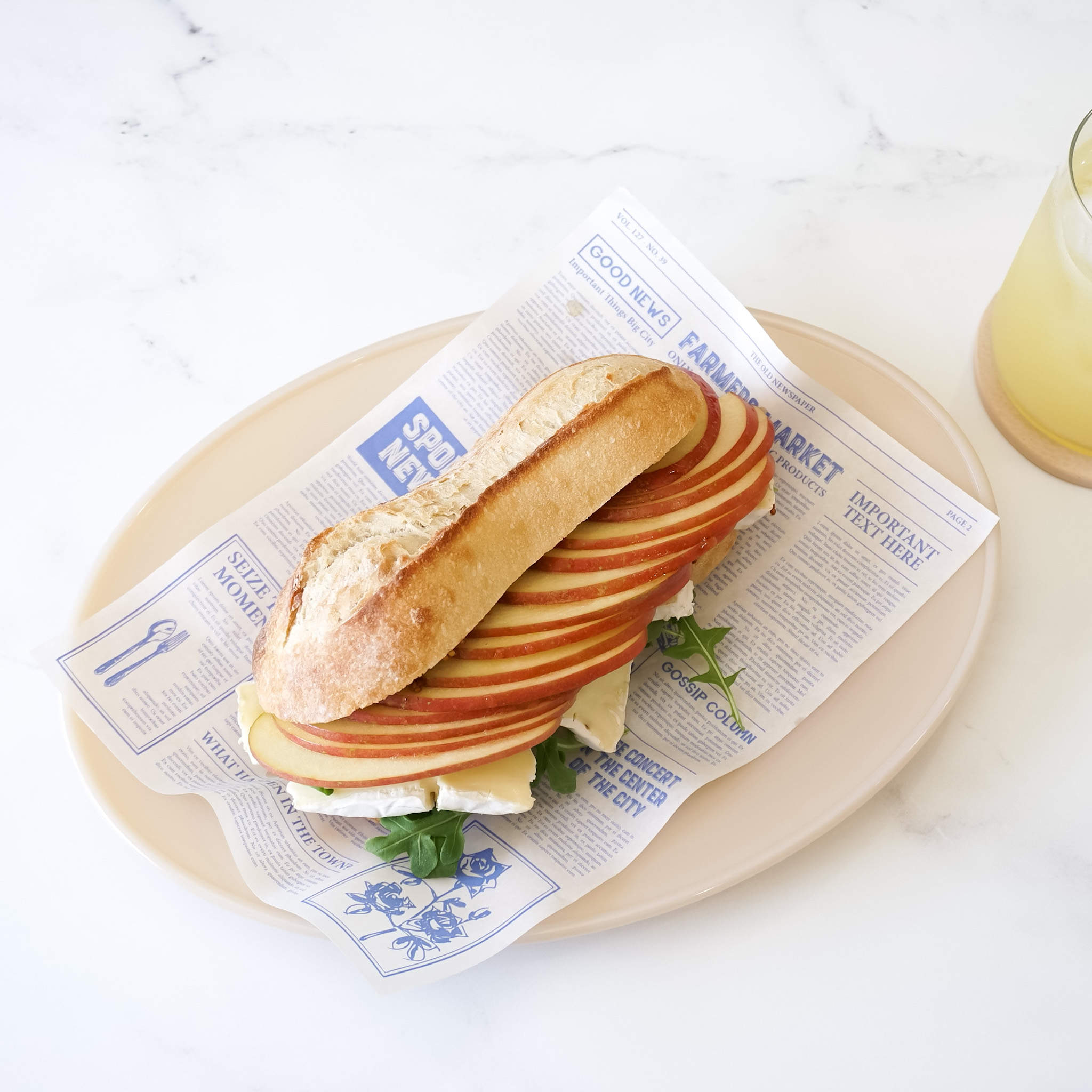 A baguette sandwich with thinly sliced red apples and a white cheese filling on a beige oval plate. The sandwich is wrapped in white news paper with blue text.
