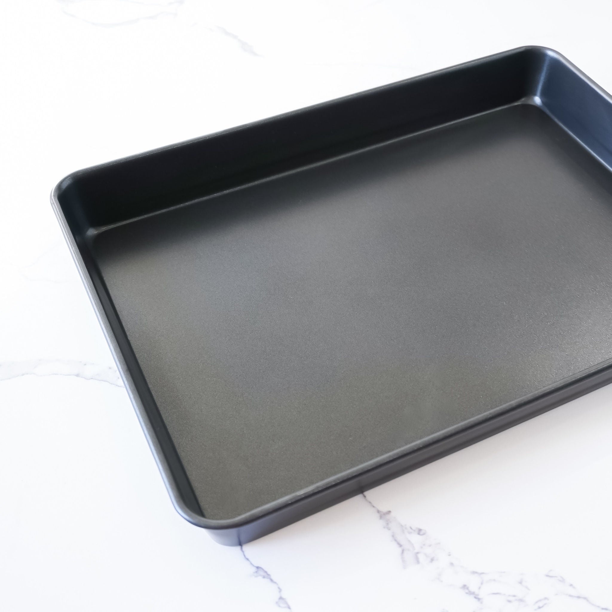 A rectangular, black sheet cake pan made of nonstick coating material, placed on a white marble surface.