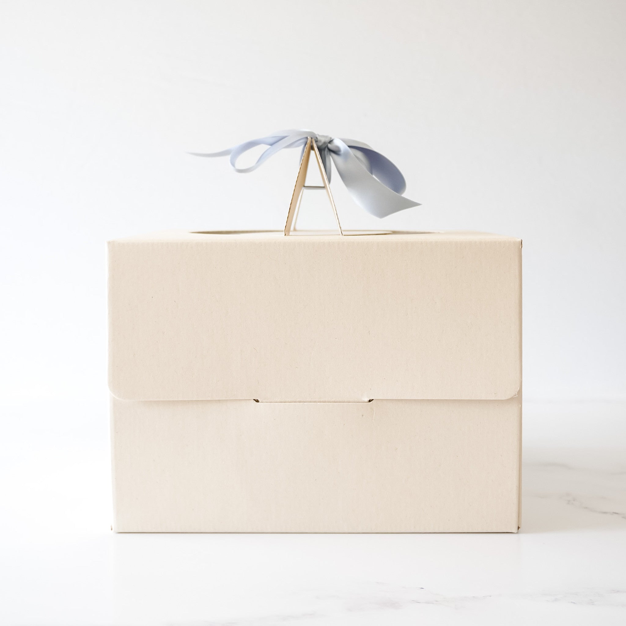 A closed, beige cake box with a light blue ribbon tied around the top. The box has a handle on the top.