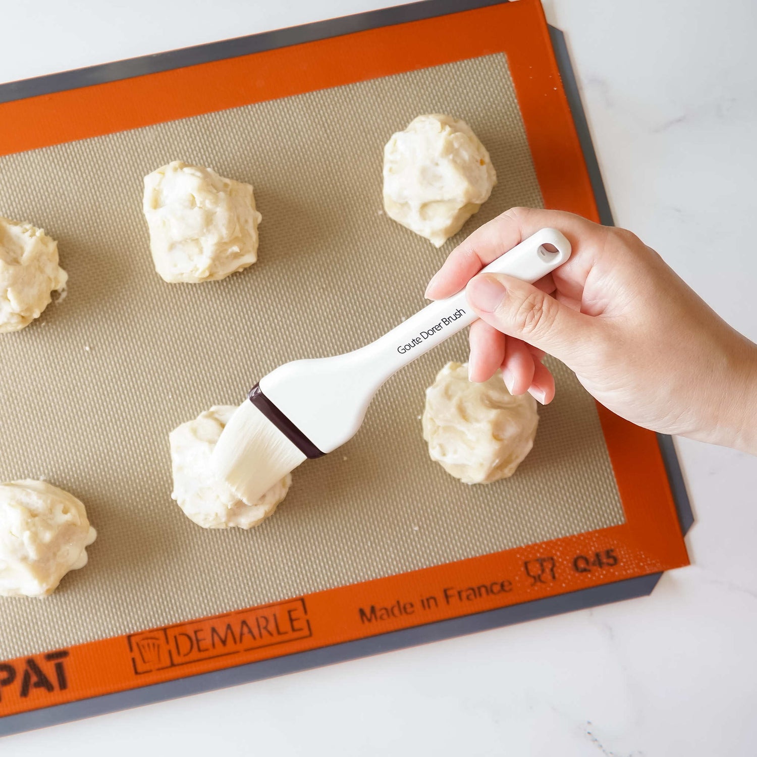 A hand using a silicone pastry brush to apply glaze to cookie dough on a silicone baking mat.