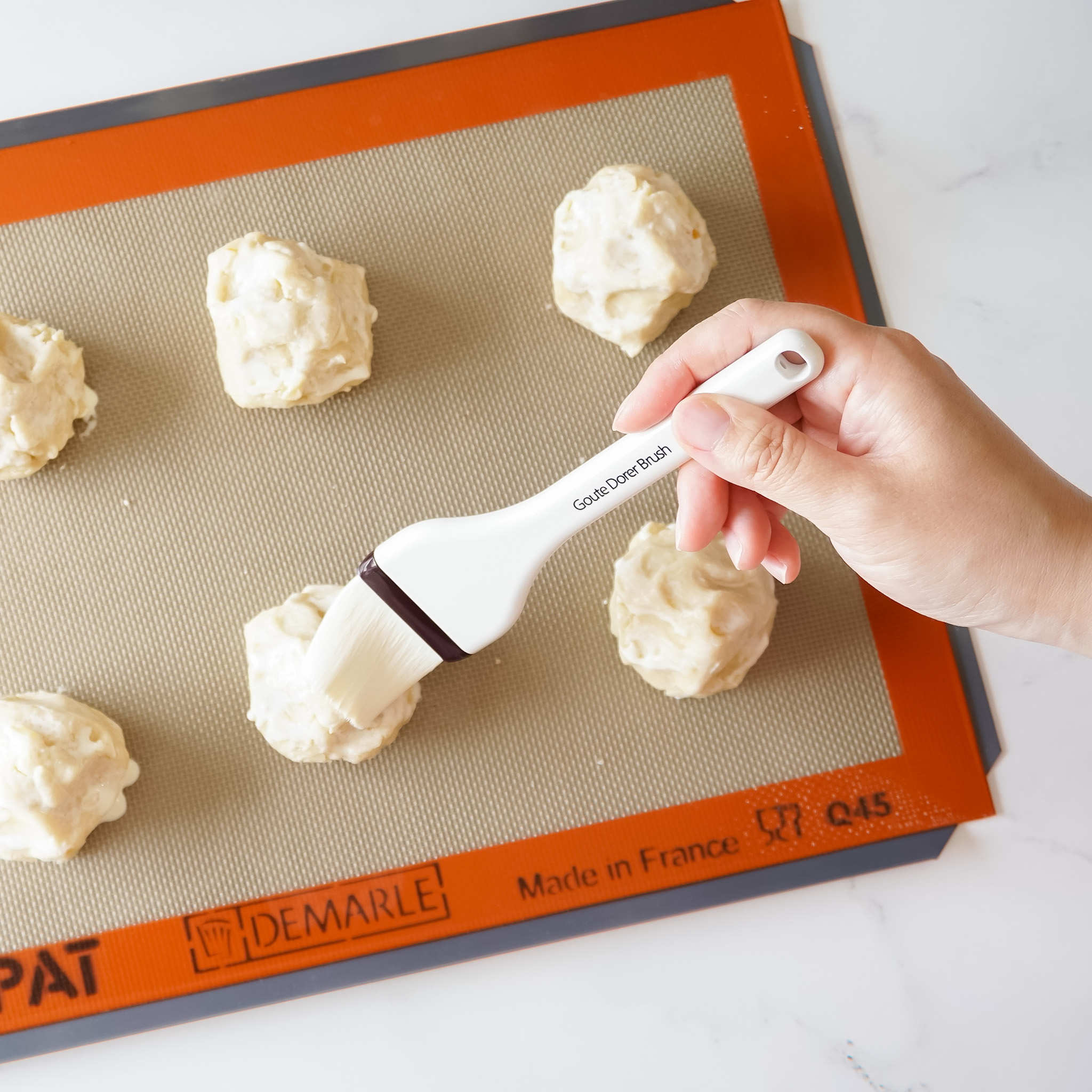 A hand using a silicone pastry brush to apply glaze to cookie dough on a silicone baking mat.