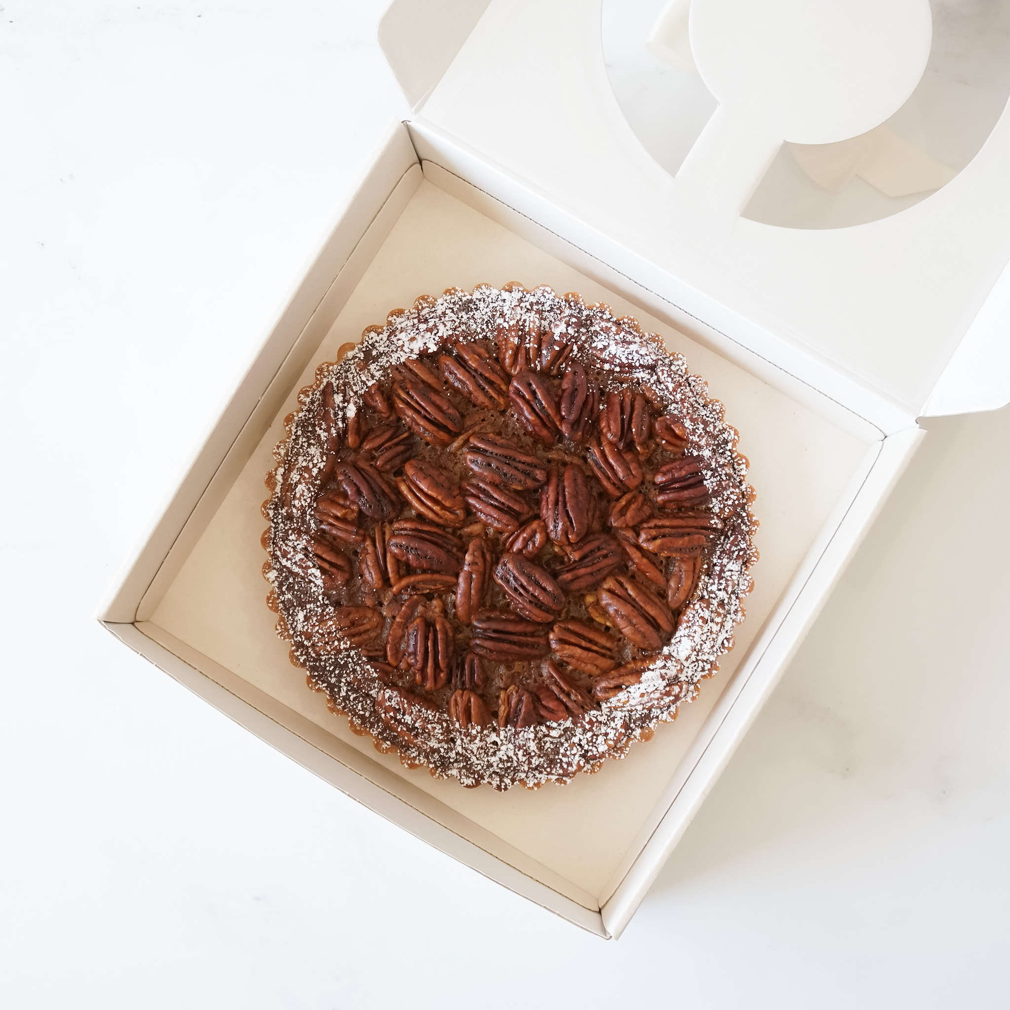 A pecan pie in a light beige cardboard box. The pie is topped with pecans and powdered sugar.