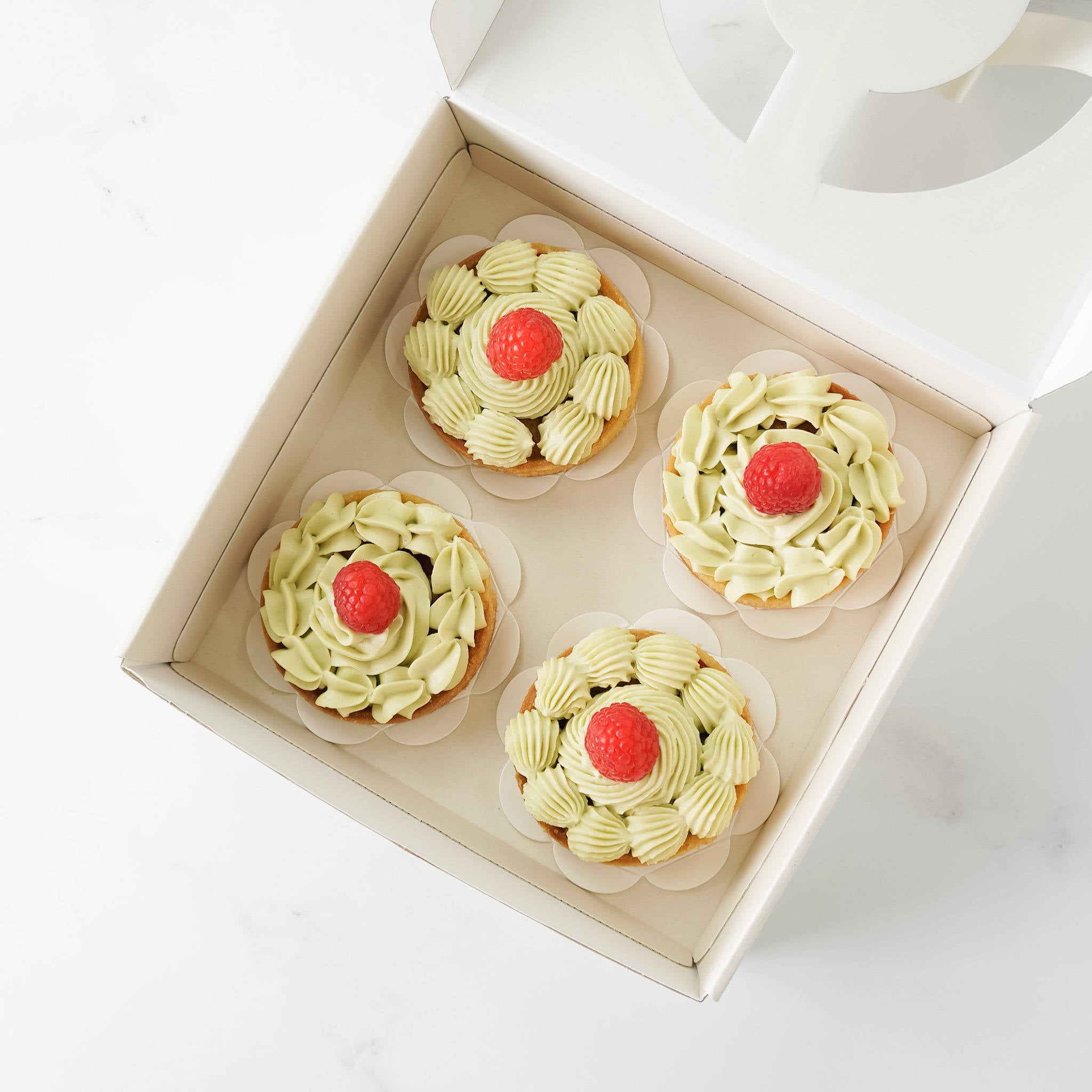 Four individual green and white mini tarts with a single red raspberry on top in a white box