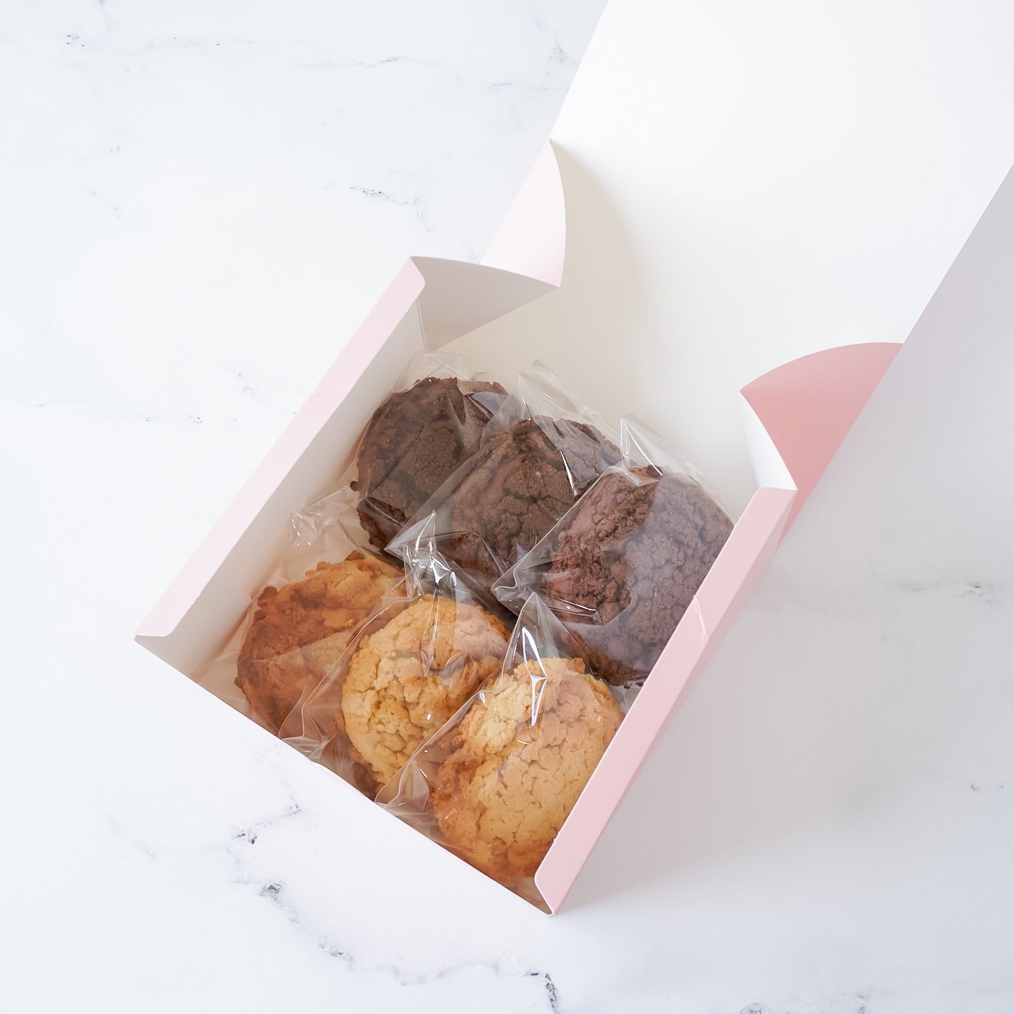 A pink box with a white interior lid open, revealing six cookies in clear plastic bags. Three cookies are brown and three are light brown. The cookies are arranged in two rows of three.