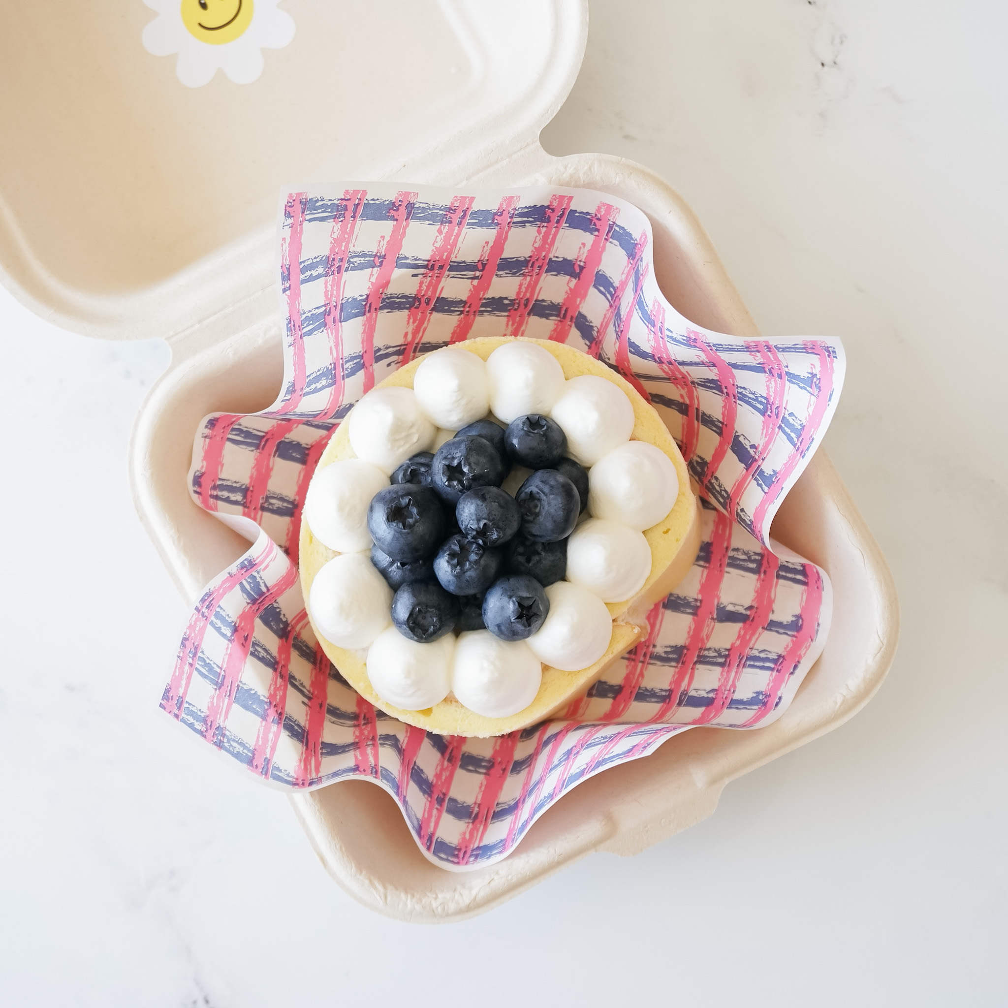 A white cake with a blue and white checkered paper liner. The cake is topped with whipped cream and blueberries.