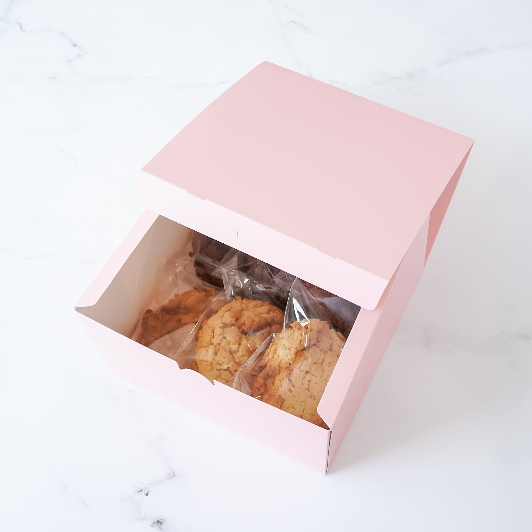 A pink box with a lid open, revealing three cookies and a brownie wrapped in plastic inside. The cookies are light brown and the brownie is dark brown.