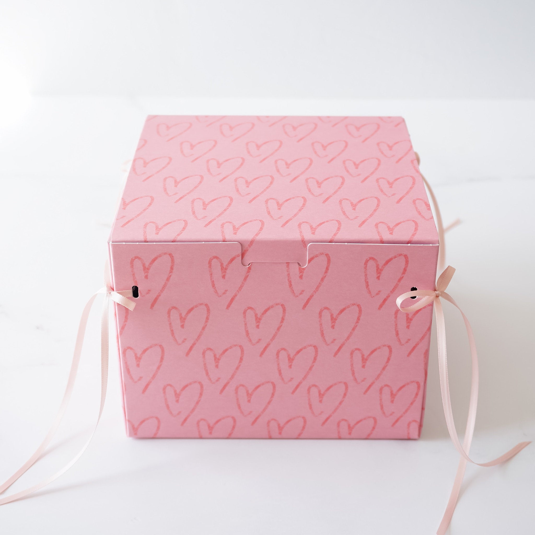 A pink square box with a heart pattern. The box has two pink ribbons attached to the sides.