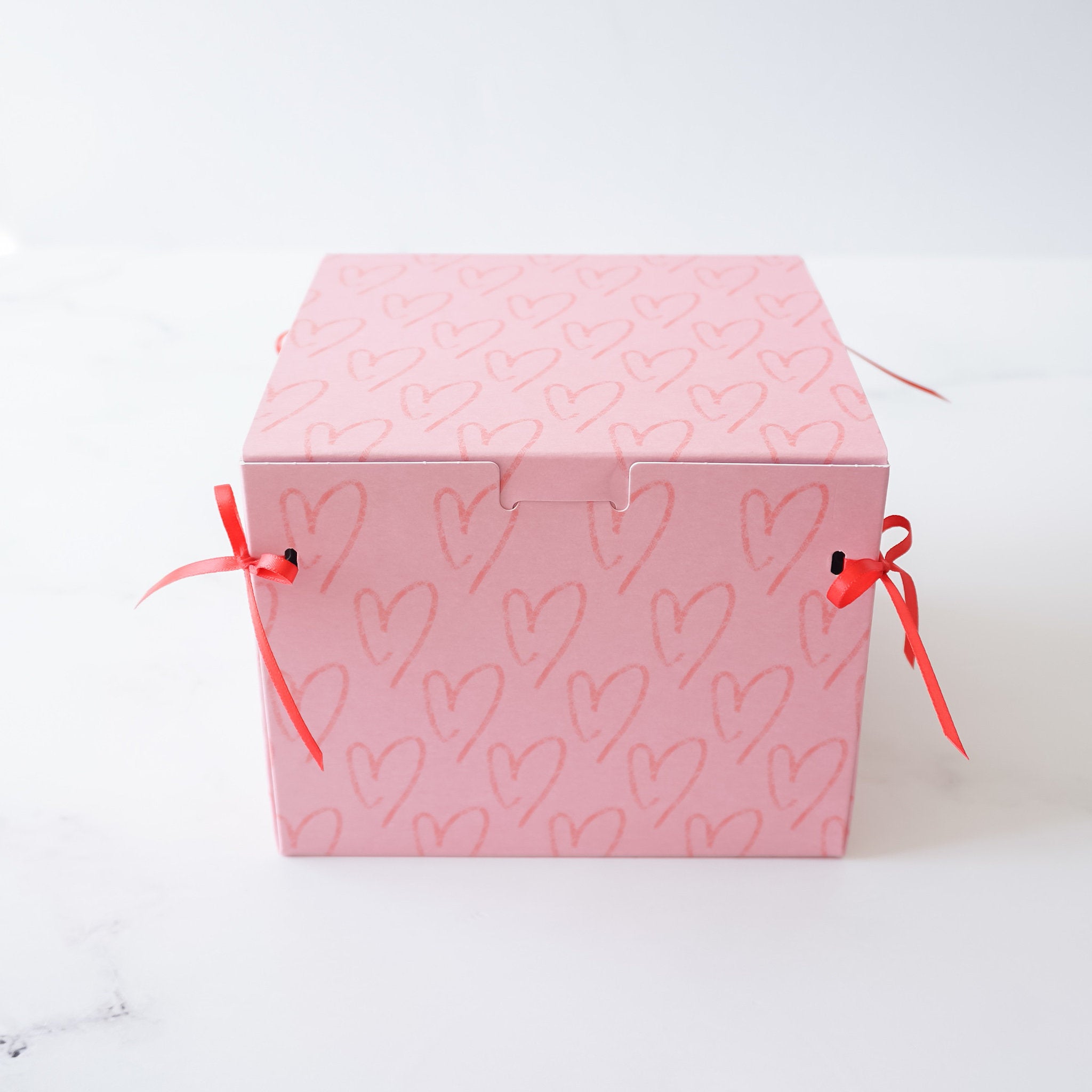 A pink gift box with a heart pattern and red ribbon ties. The box is closed and ready for gifting.