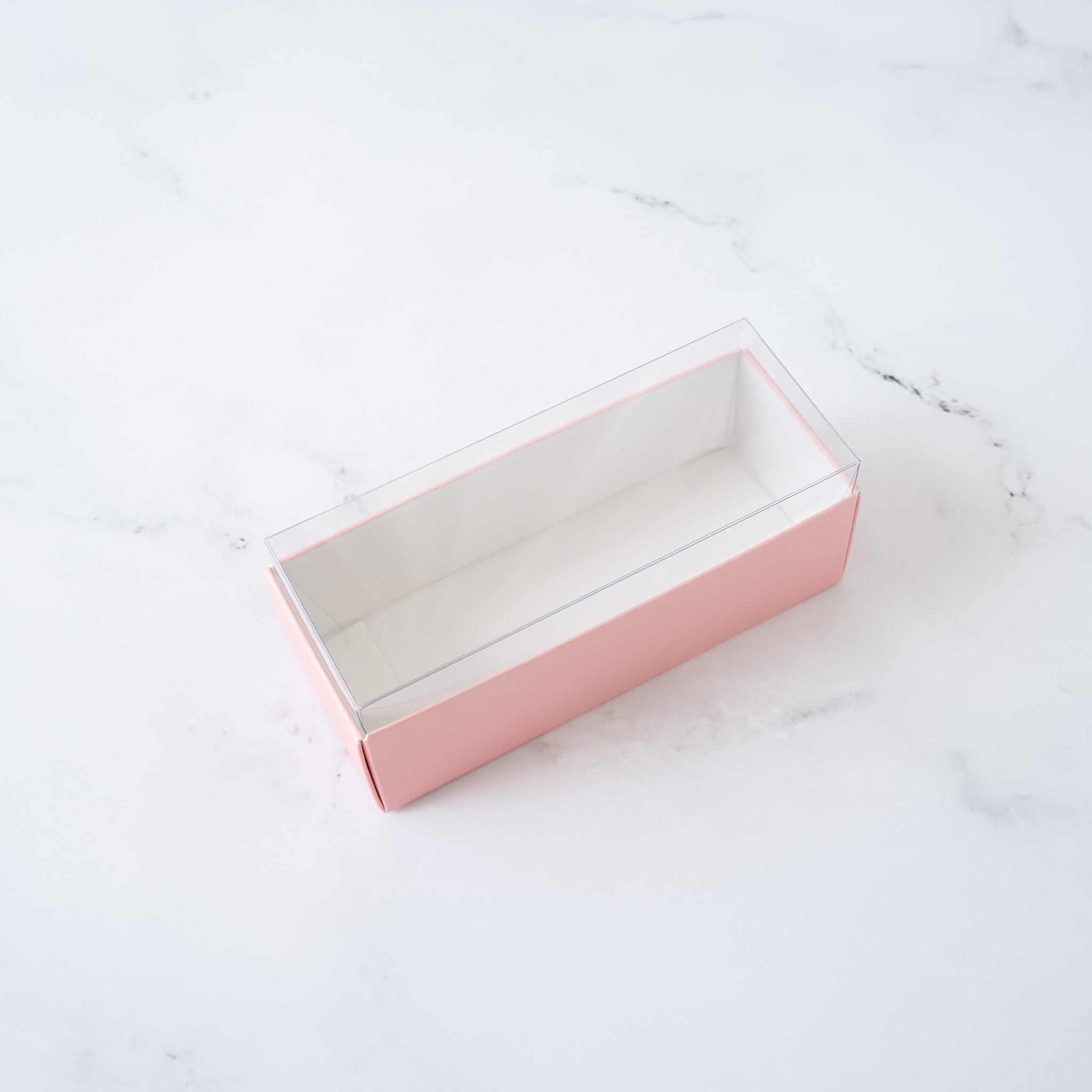 A pink cardboard box with a clear plastic lid. The box is empty and is sitting on a white marble surface.