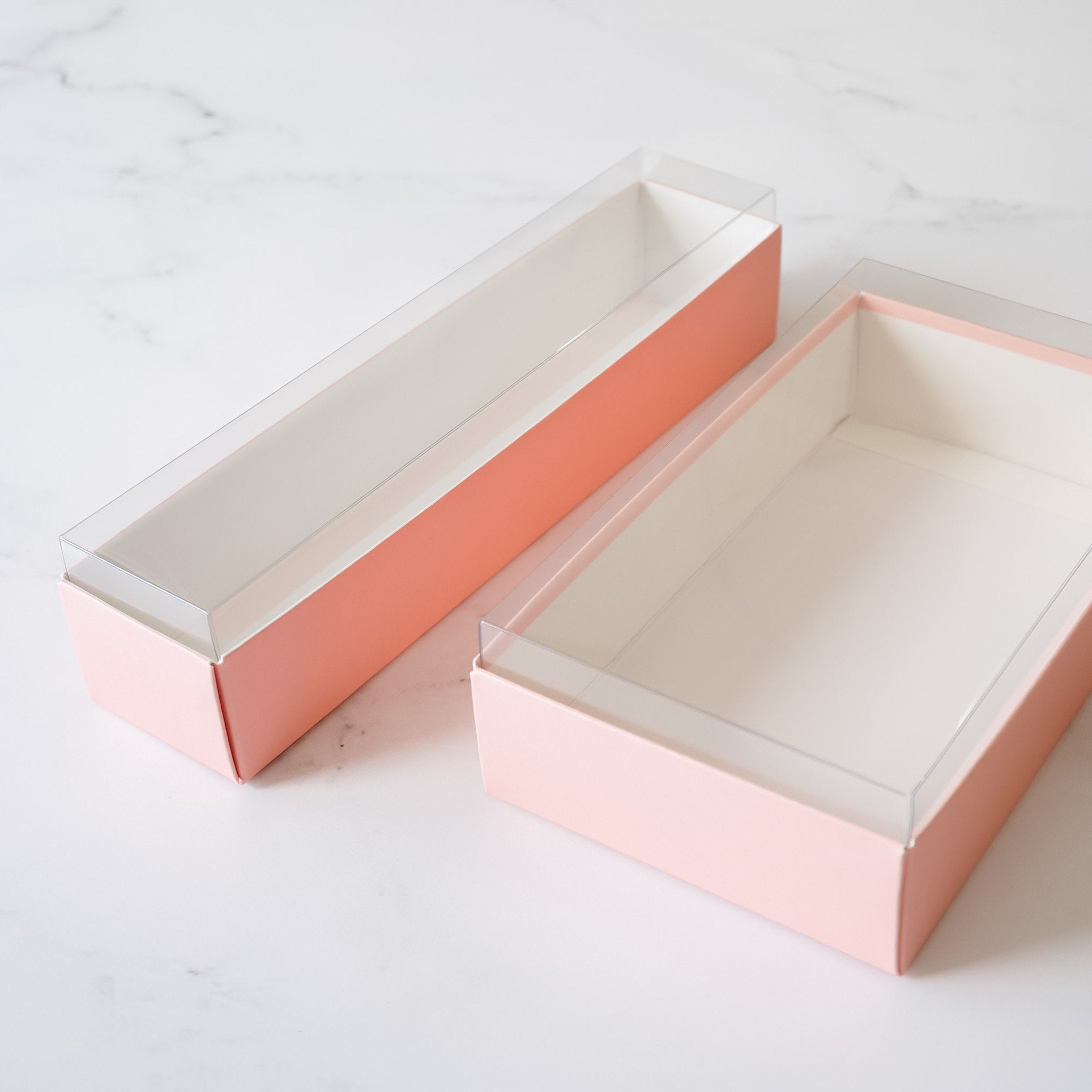 Two pink rectangular boxes with clear plastic lids. The boxes are empty and are sitting on a white marble surface.