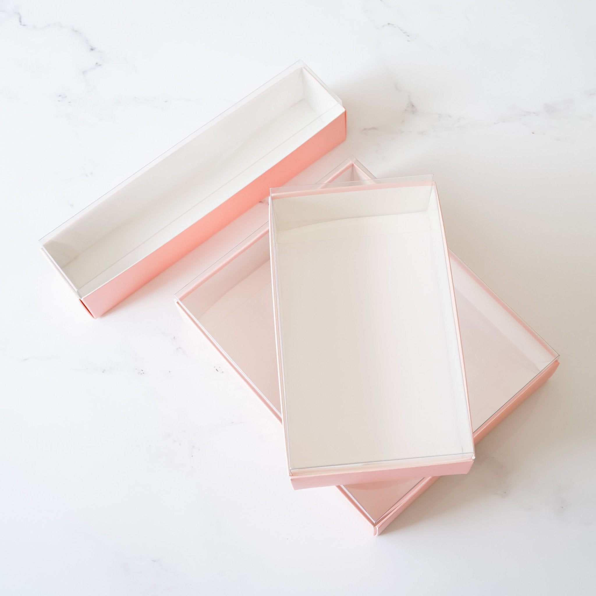 Three pink gift boxes with clear lids. The boxes are stacked on top of each other. The top box is open and the lid is lying next to it.