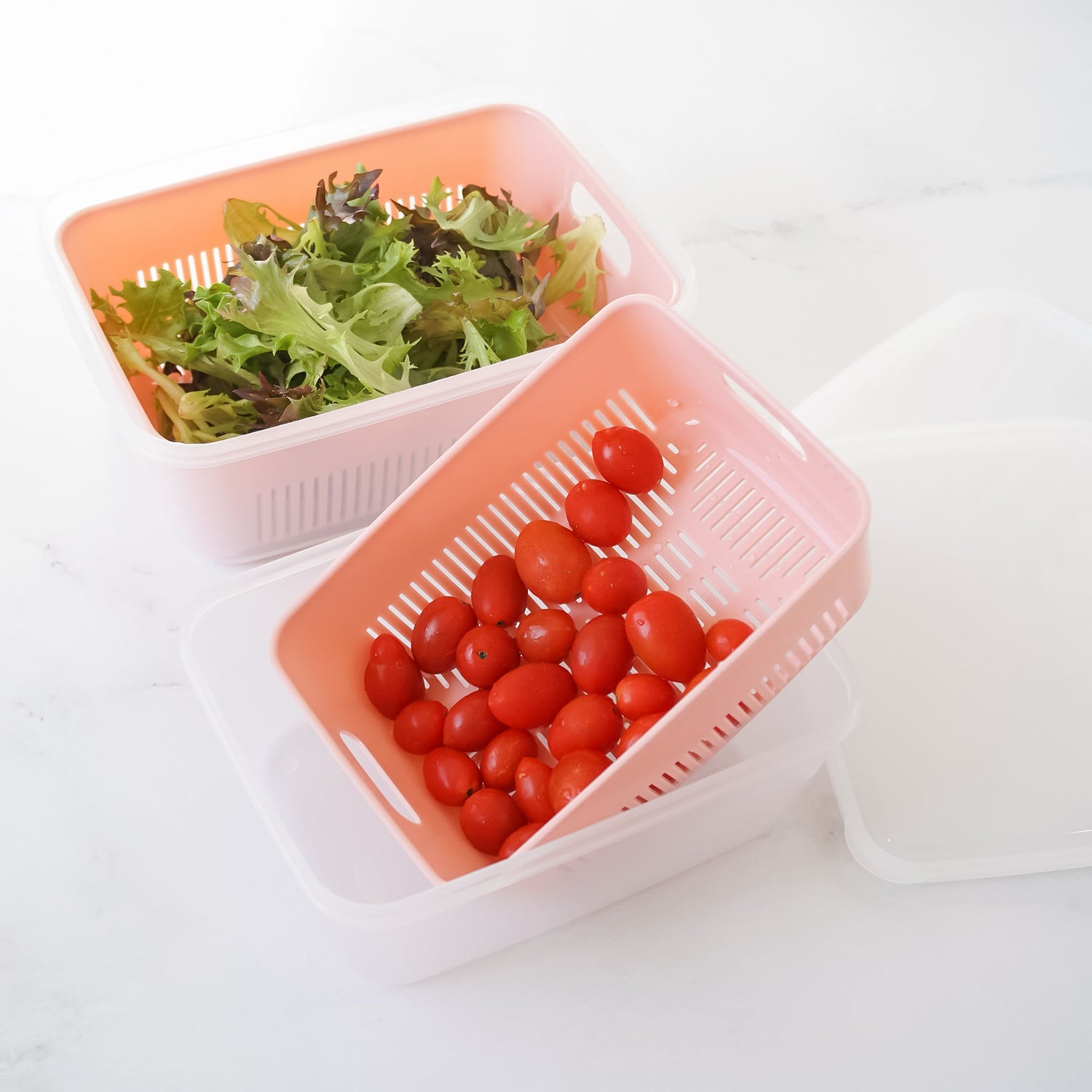 Two plastic fruit containers with one containing tomatoes and the other with salad leaves. Both are in a pink color with a white lid placed beside them.