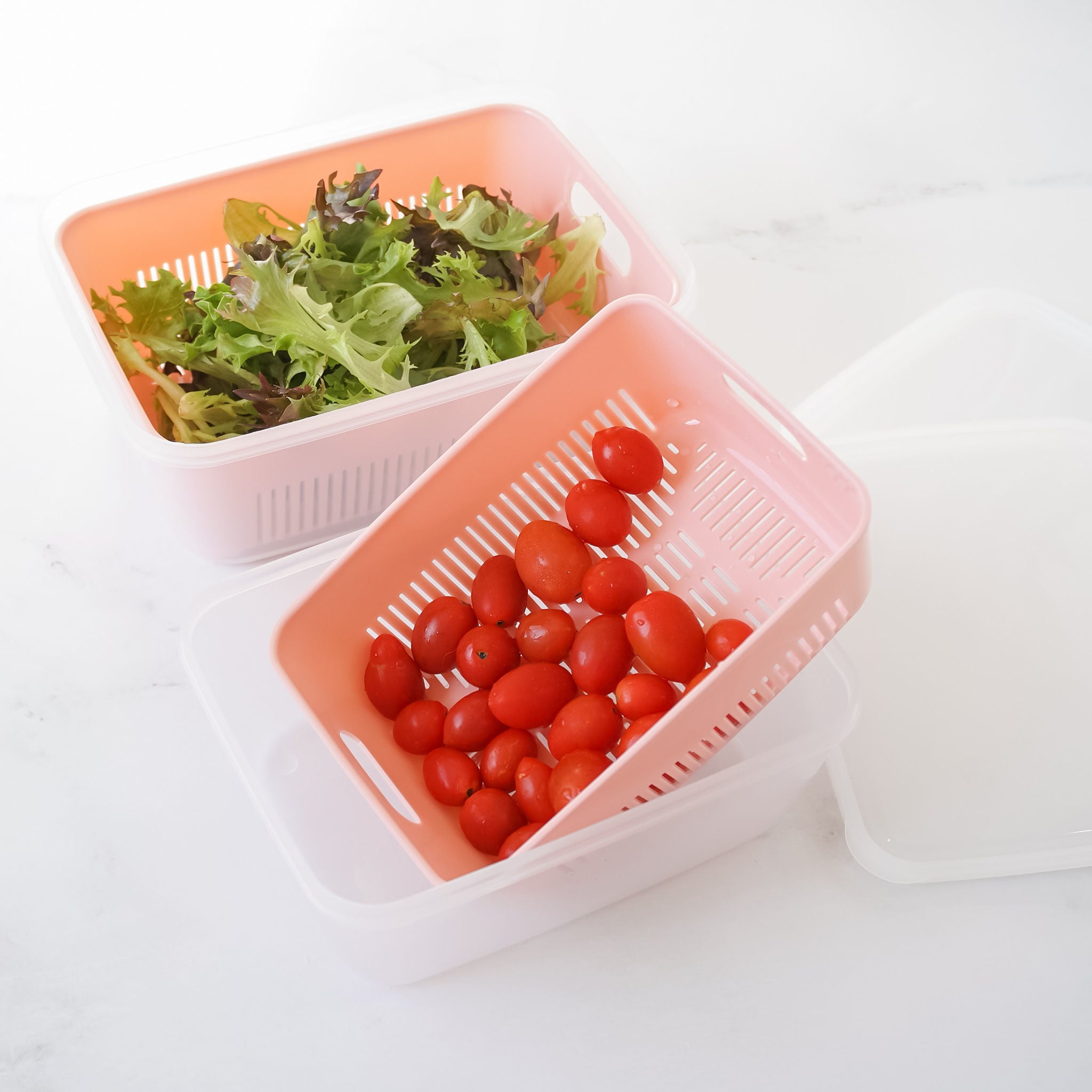 Two plastic fruit containers with one containing tomatoes and the other with salad leaves. Both are in a pink color with a white lid placed beside them.