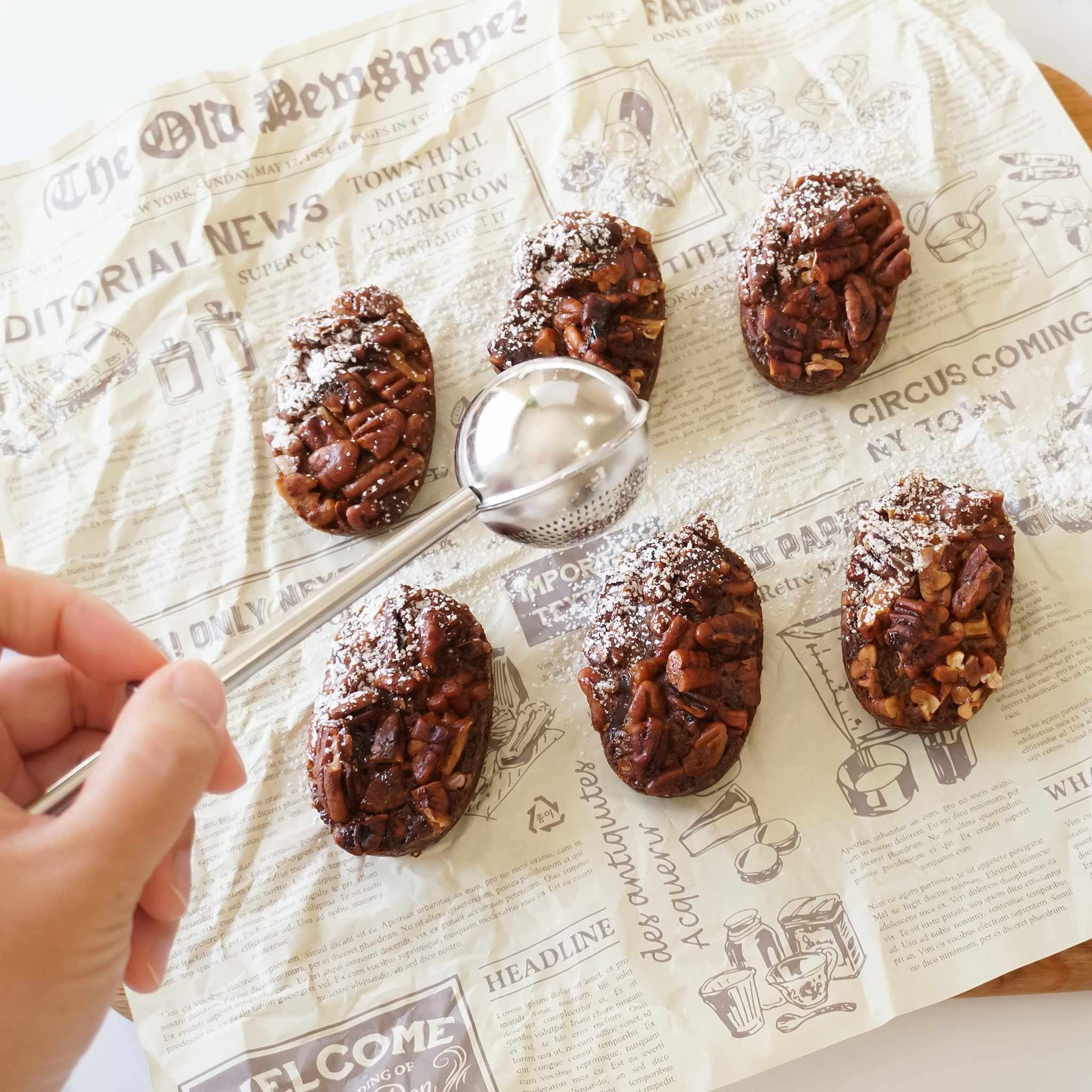 A powdered sugar shaker made of stainless steel, shown with a perforated section for dusting powdered sugar and a solid section for scooping sugar or flour, positioned on a wooden surface with a piece of paper that has printed text.