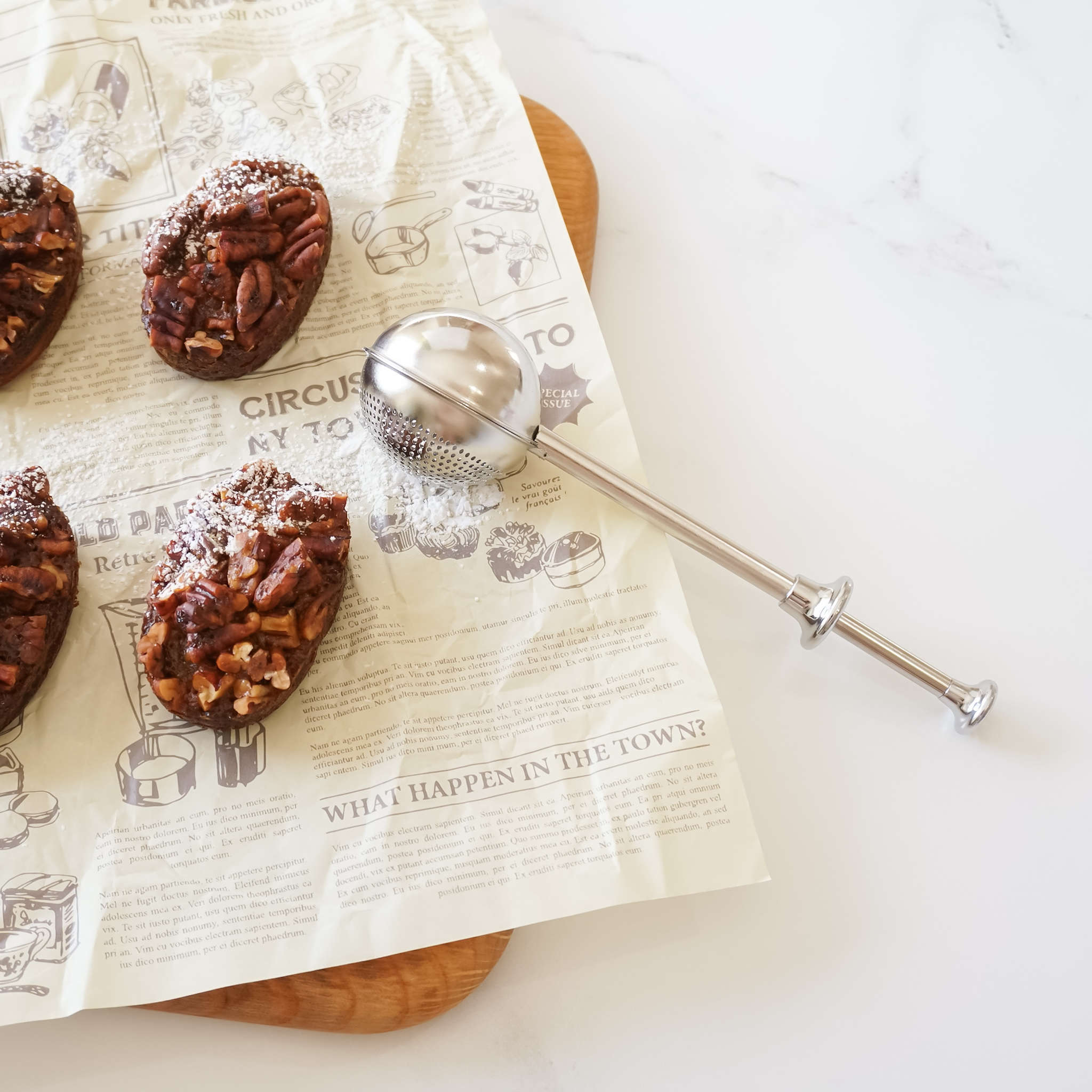 A powdered sugar shaker made of stainless steel, shown with a perforated section for dusting powdered sugar and a solid section for scooping sugar or flour, positioned on a wooden surface with a piece of paper that has printed text.