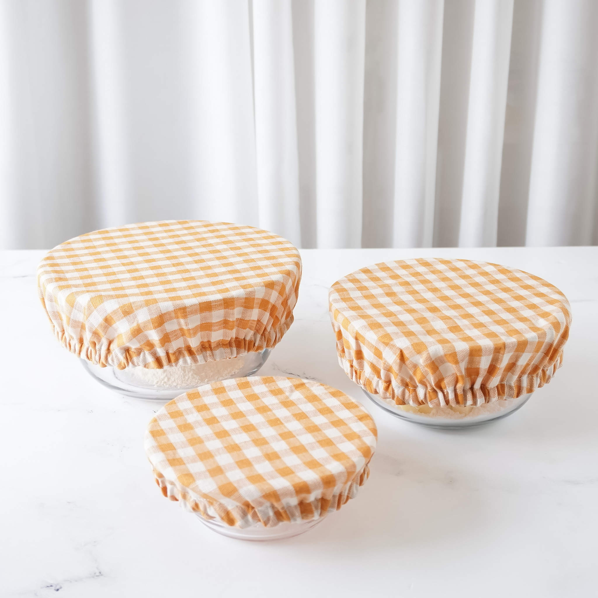 Three bowl covers in orange and white checkered pattern, sized small, medium, and large, displayed on a white surface with a draped white curtain in the background.