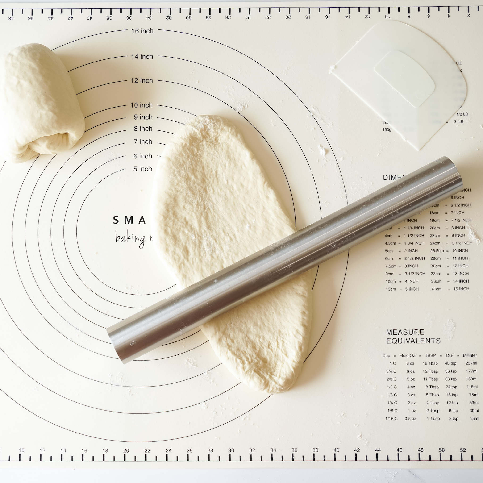 A stainless steel rolling pin is displayed on a silicone baking mat with a rolled out piece of dough beside it, along with a measuring guide.