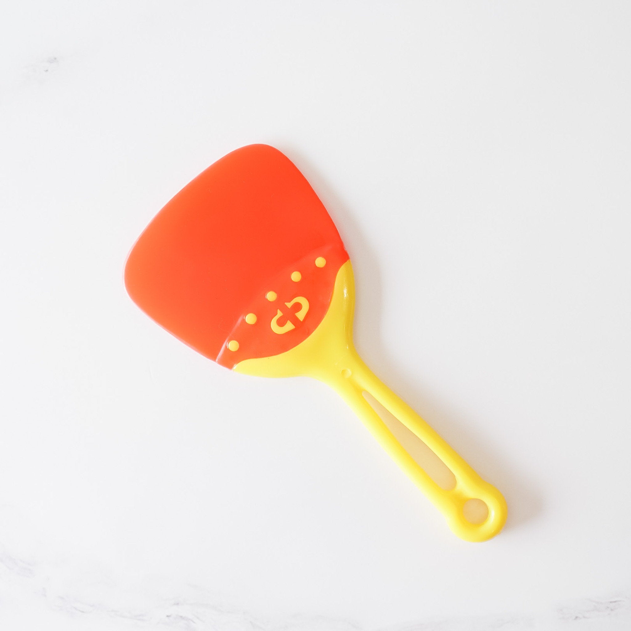 A yellow and red plastic spatula with a heart-shaped logo. The spatula has a slightly curved, wide blade and a long, slender handle. The handle has a small hole at the end for hanging. The spatula is designed for cooking and food preparation.