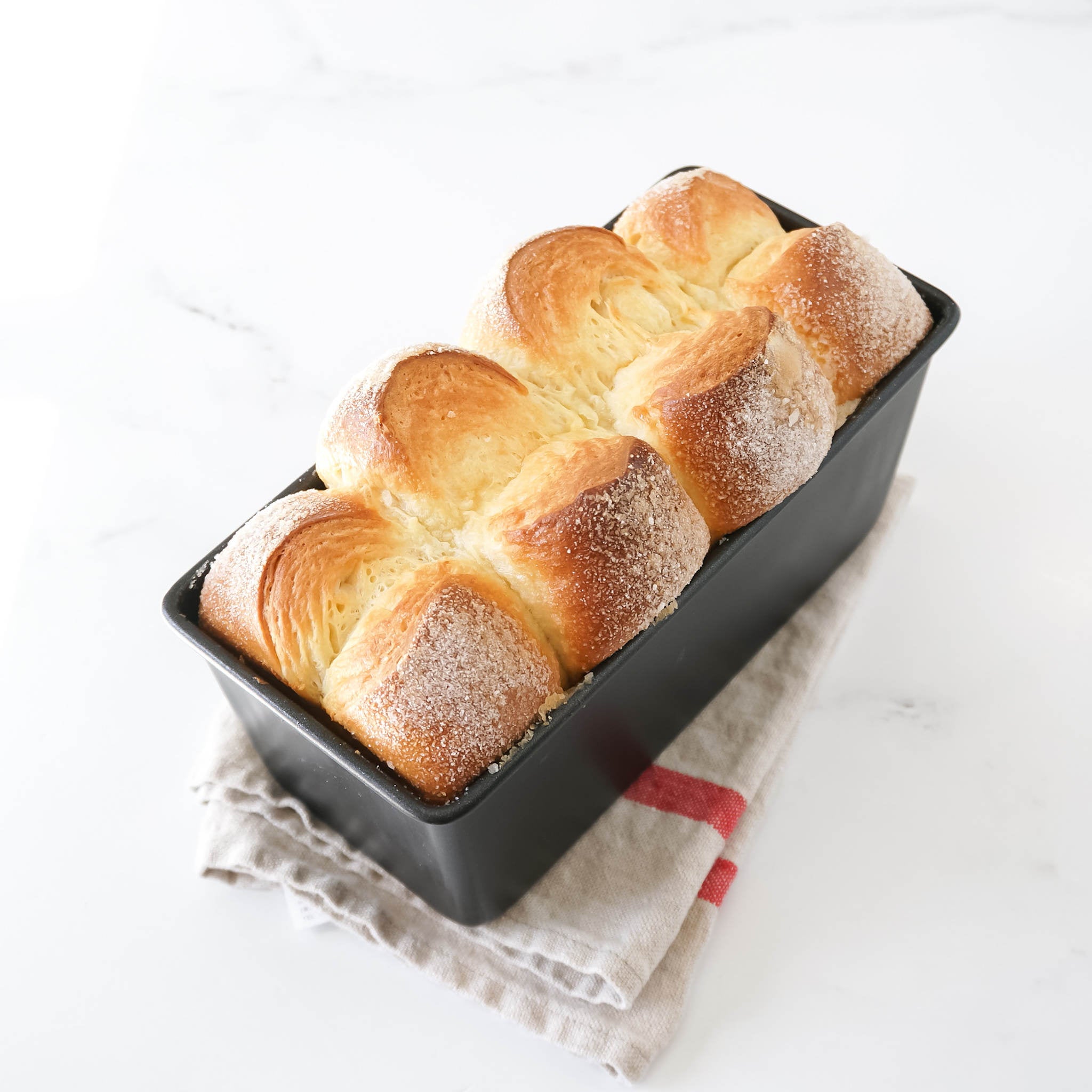 A freshly baked loaf of bread in a black bread loaf pan, placed on a white surface with a red and white cloth to the side.