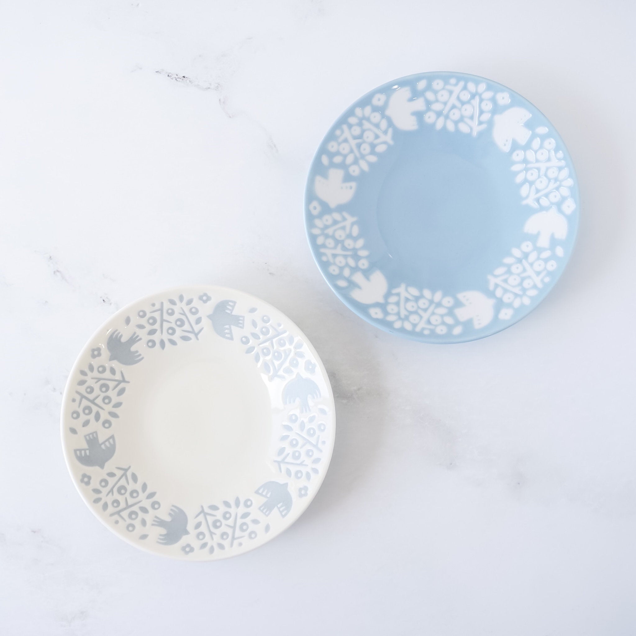 Two Mino Ware small plates with embossed patterns of birds and plants, one in white and one in blue, displayed on a marble surface.