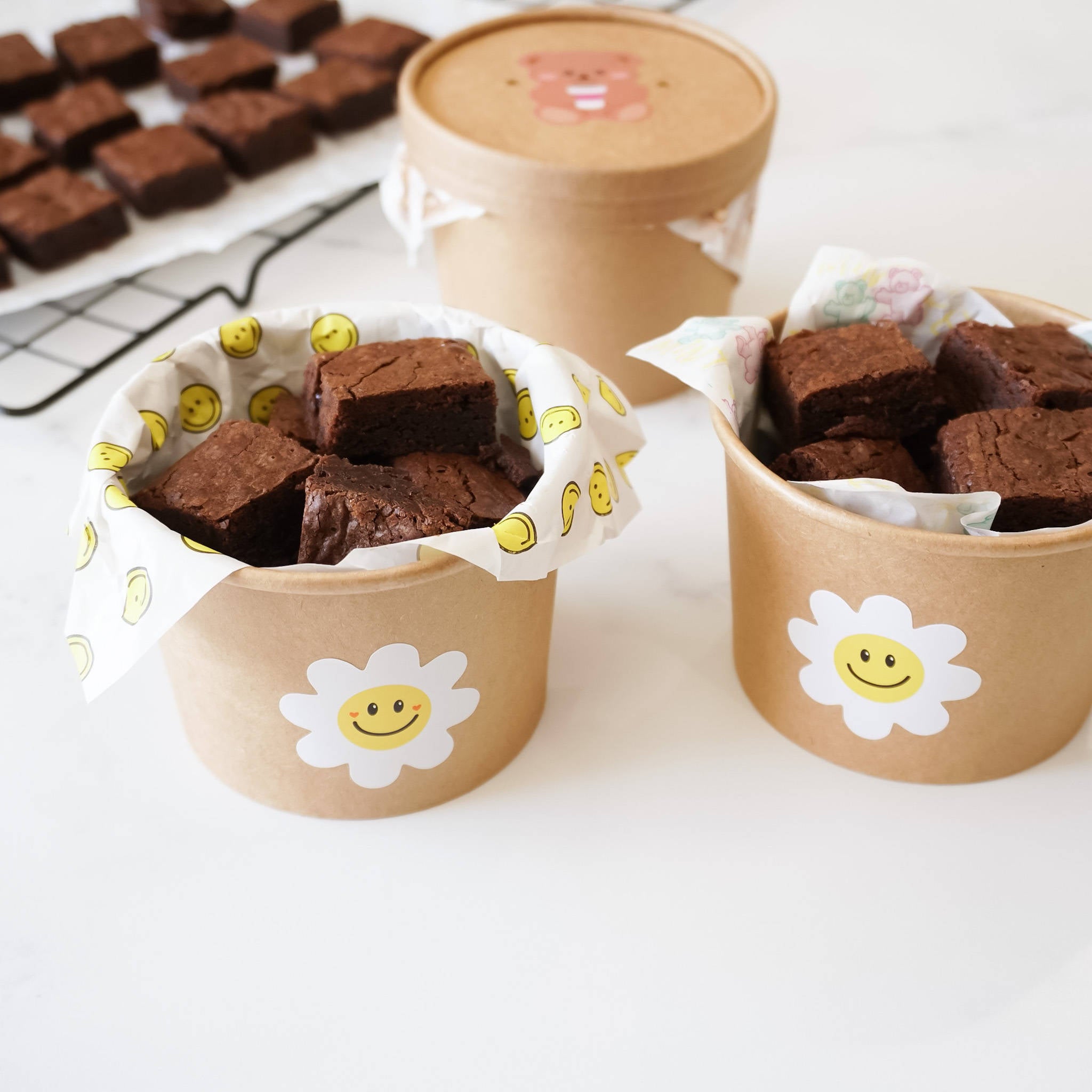 Two brown paper cups with white daisy stickers with yellow smiley faces. The cups are filled with chocolate brownies and lined with white paper with a yellow smiley face pattern.