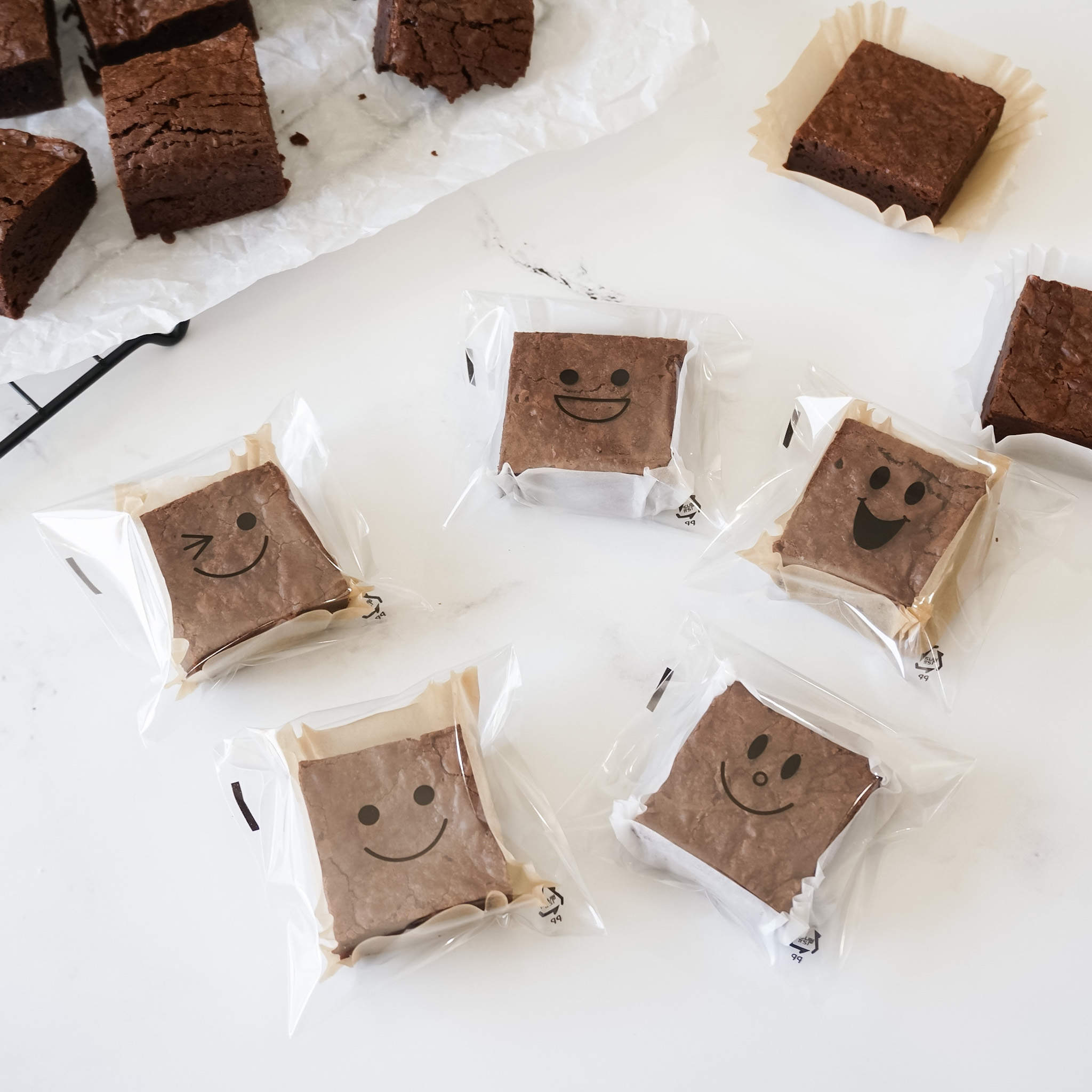 Six individually packaged chocolate brownies with different smiley faces printed on them. The brownies are wrapped in clear plastic bags and are on a white surface.