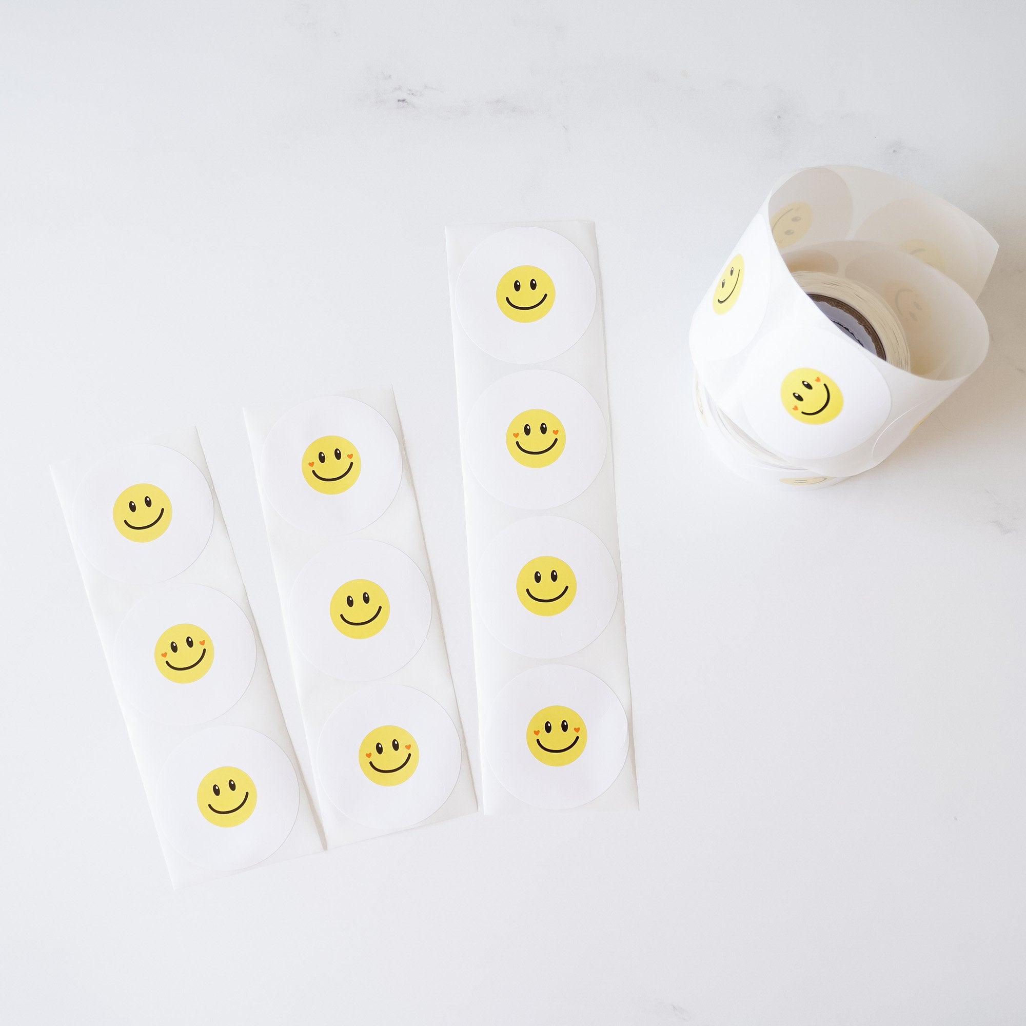Three sheets of white paper with yellow smiley face stickers. Each sheet has six stickers. The stickers have black eyes and a black smile. The stickers are arranged in a vertical row on each sheet. The sheets are on a white surface.