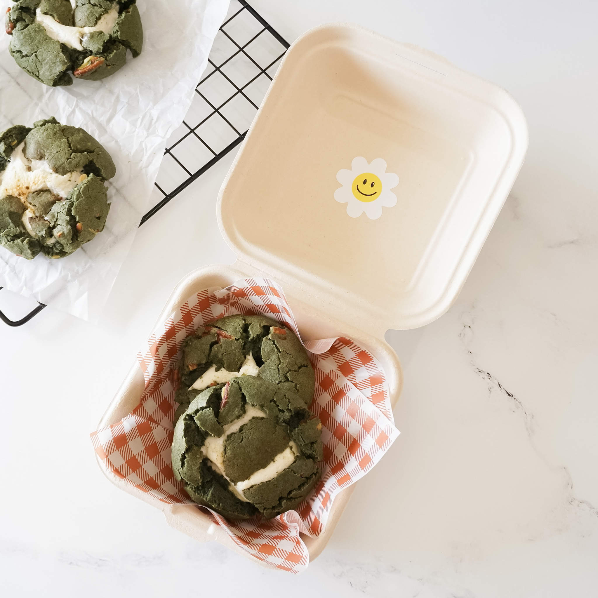 Two green and white cookies with a white swirl design are in a white cardboard container with a red and white checkered liner. The container has a smiley face daisy sticker on the top.