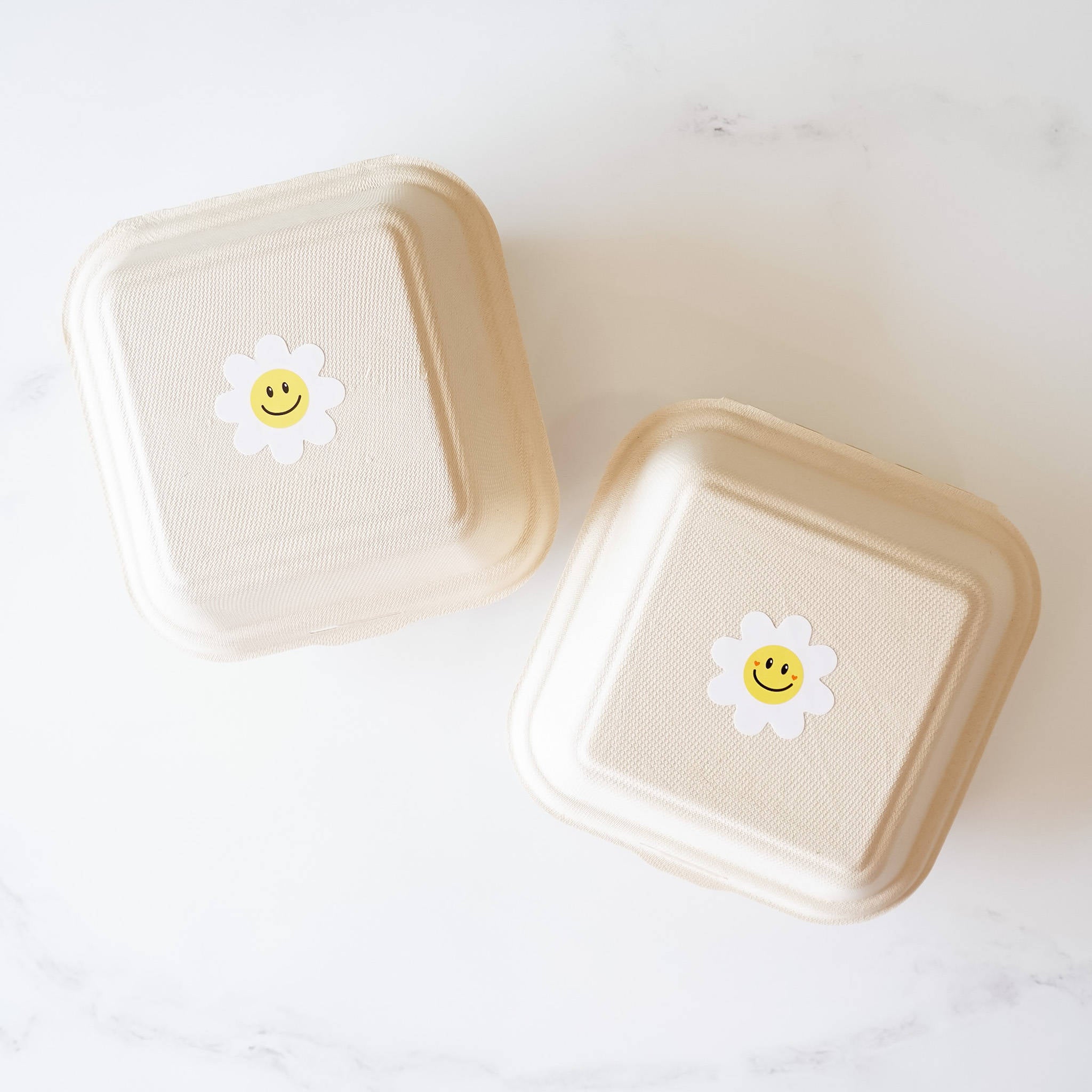 Two square, beige, cardboard takeout containers with white daisy stickers with yellow smiley faces on them.