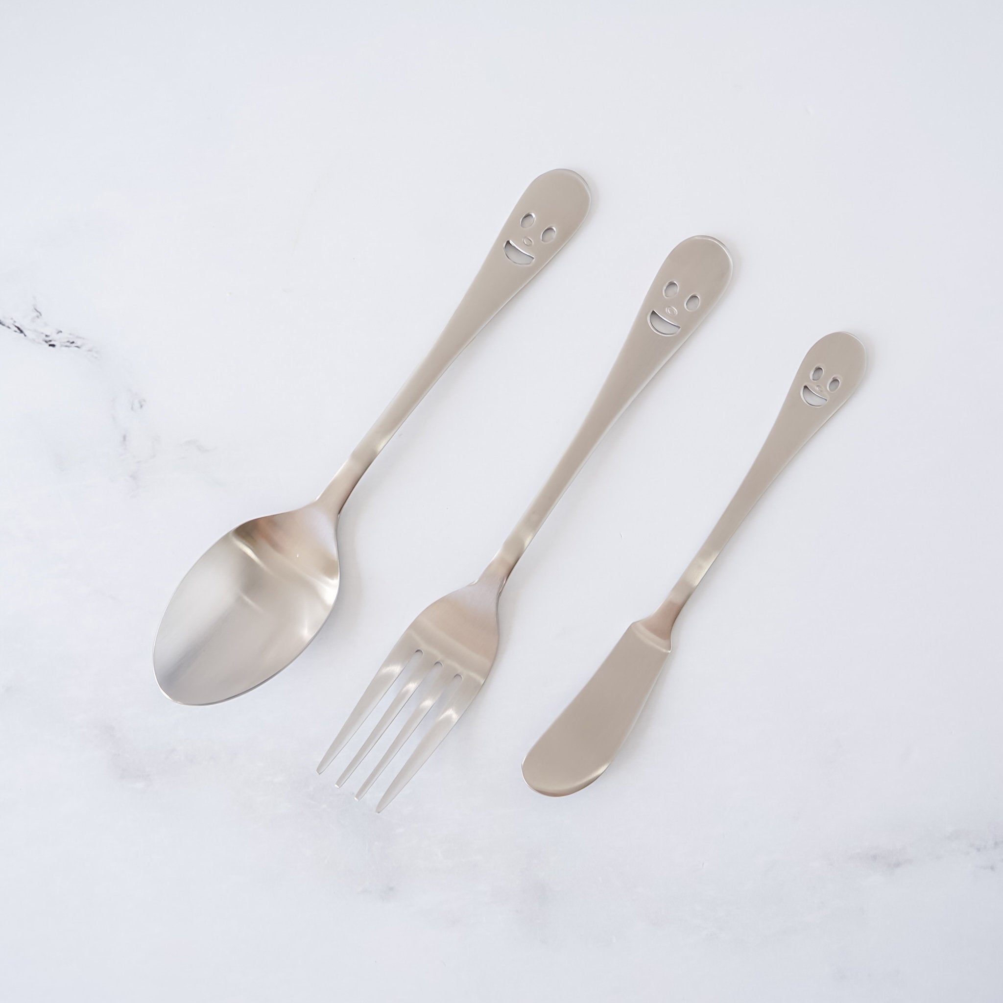 Set of three silver smiley face cutlery including a spoon, fork, and knife on a white background