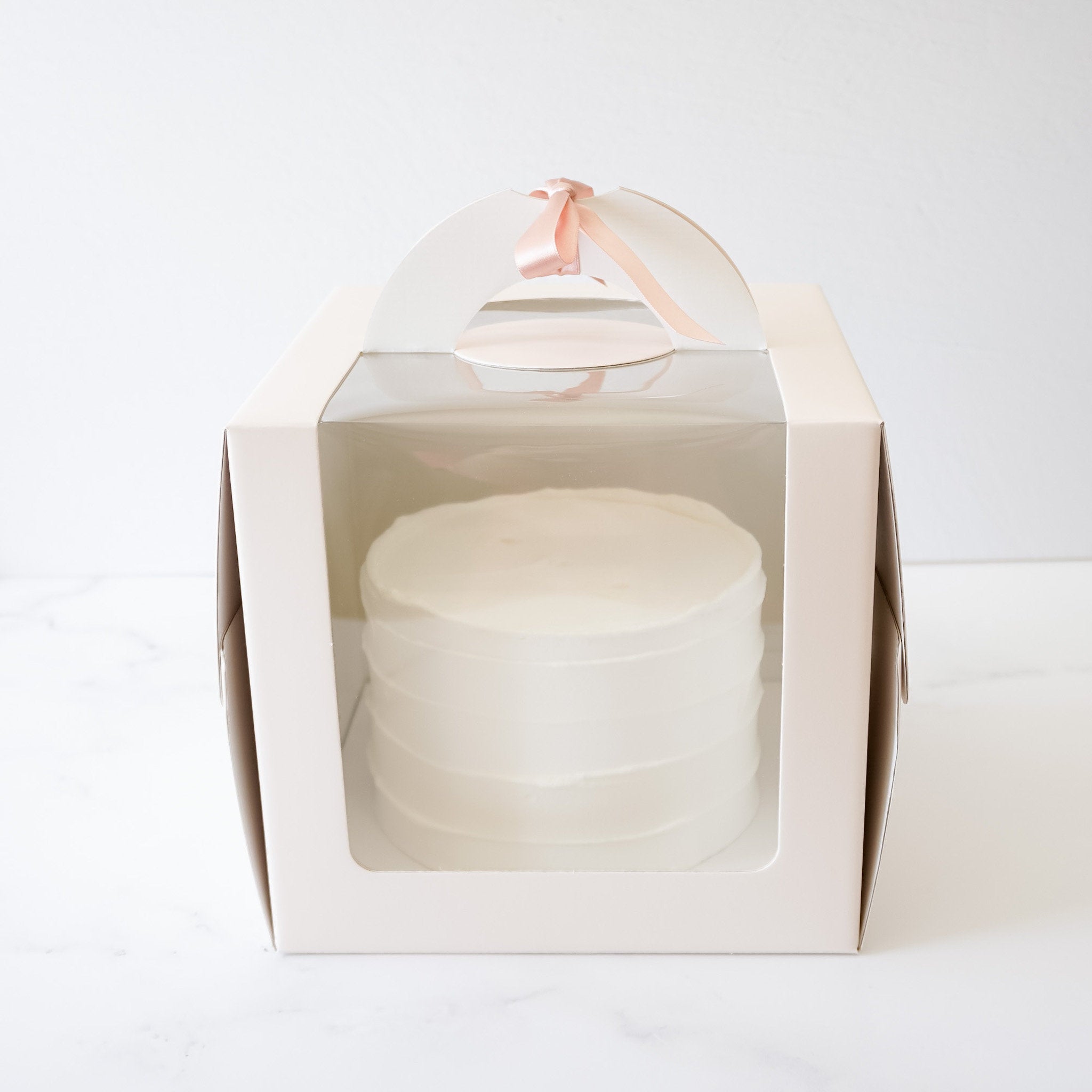A white cardboard cake box with a clear plastic window. The box has a handle with a pink ribbon tied around it. The box is designed for carrying and displaying cakes.