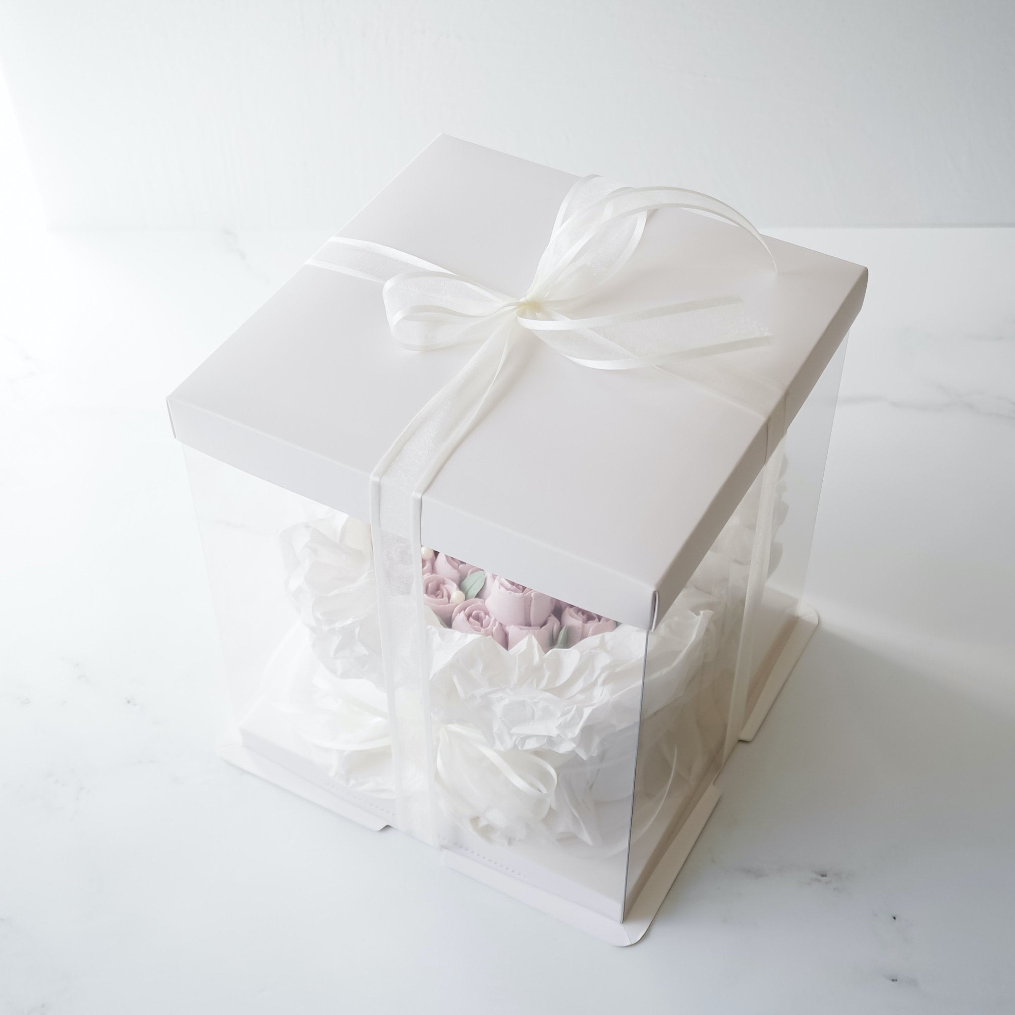 A white square box with a clear plastic window. The box is tied with a white ribbon and contains a pink and white floral arrangement.