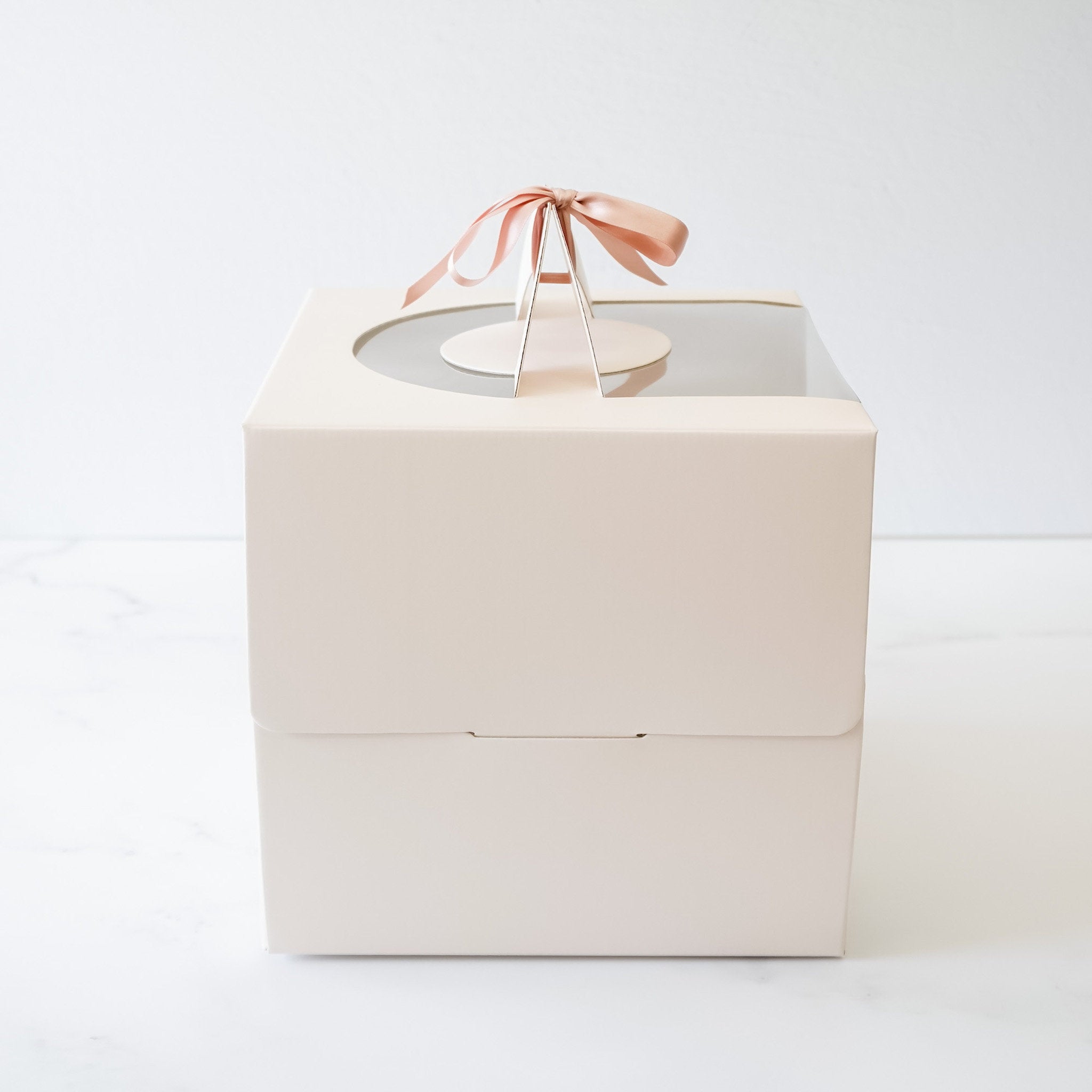 An ivory square box with a clear plastic window on the top. The box has a handle on the top with a pink ribbon tied around it. The box is designed to hold a cake or other baked goods.