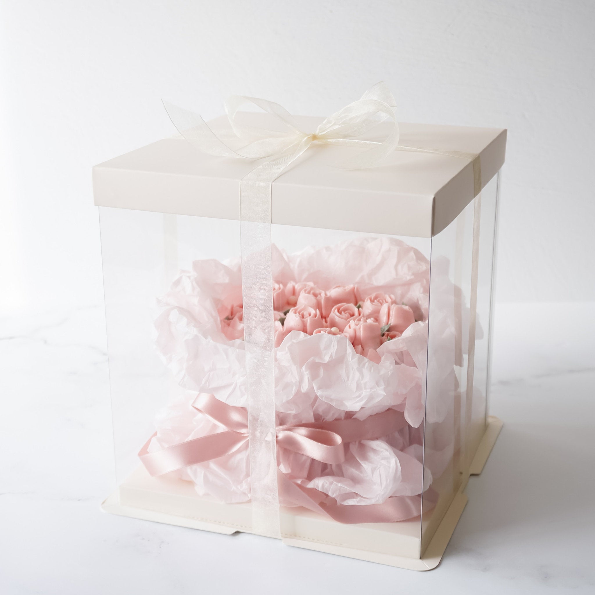 A pink rose bouquet cake wrapped in white tissue paper and tied with a pink ribbon. The bouquet is sitting on a white cardboard box with a clear acrylic lid.