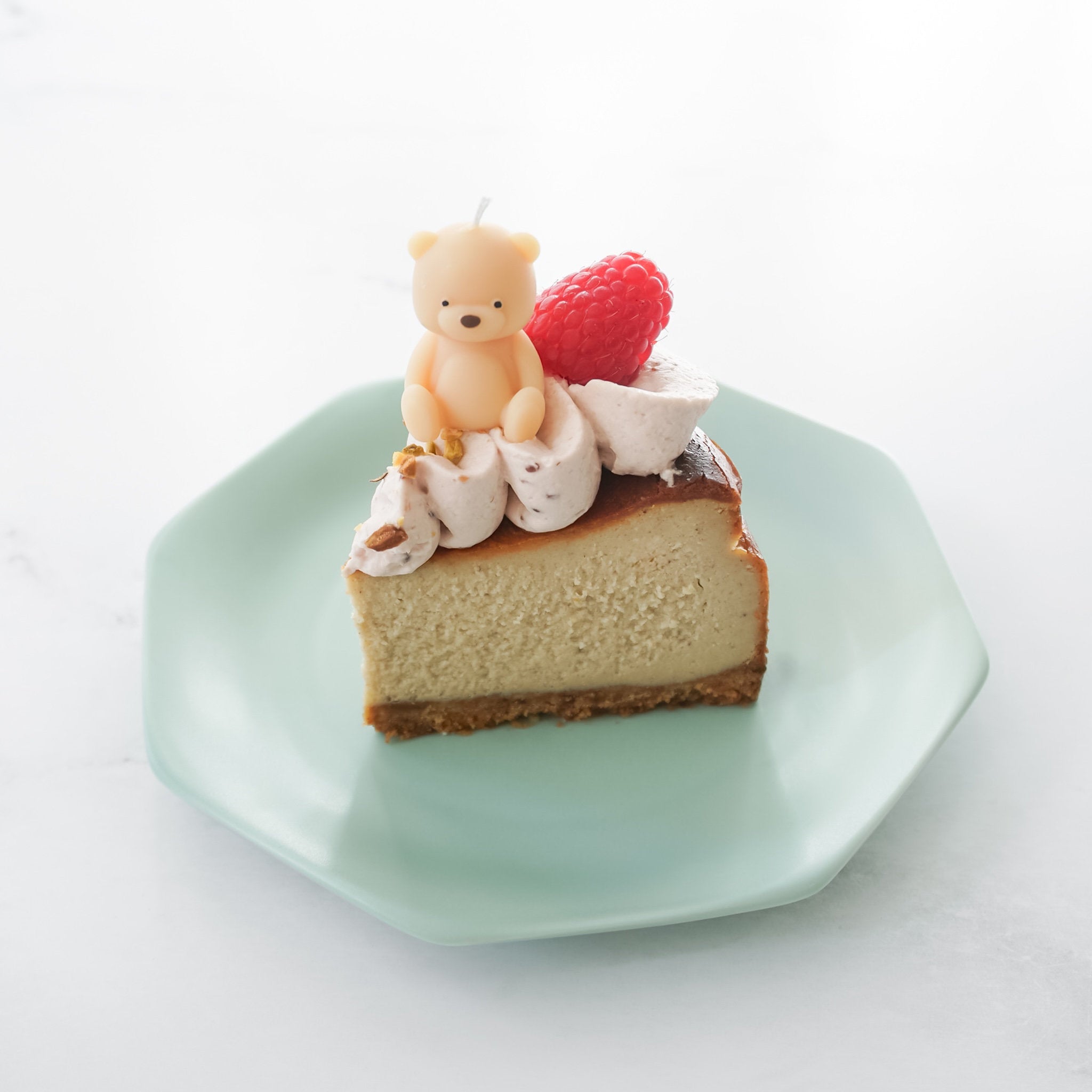Slice of cheesecake with a teddy bear and raspberry on top, served on a green plate.