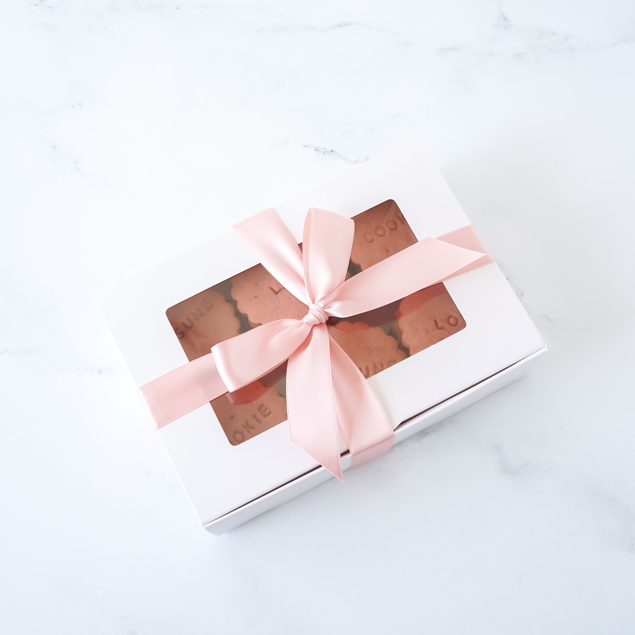 A white cookie box with a clear window on top, decorated with a pink ribbon.