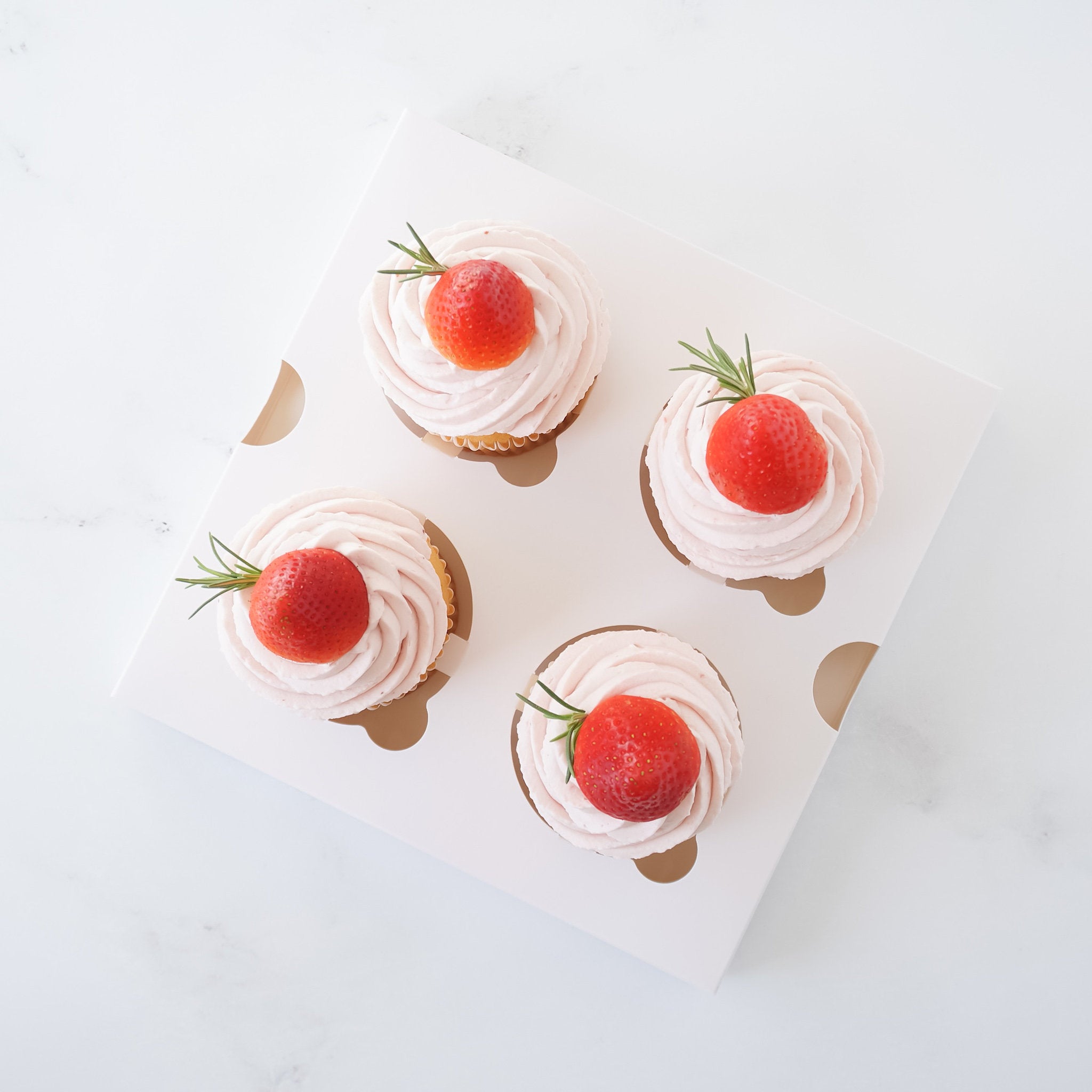 Four cupcakes with white frosting and a strawberry on top, presented in a glossy white cupcake insert.