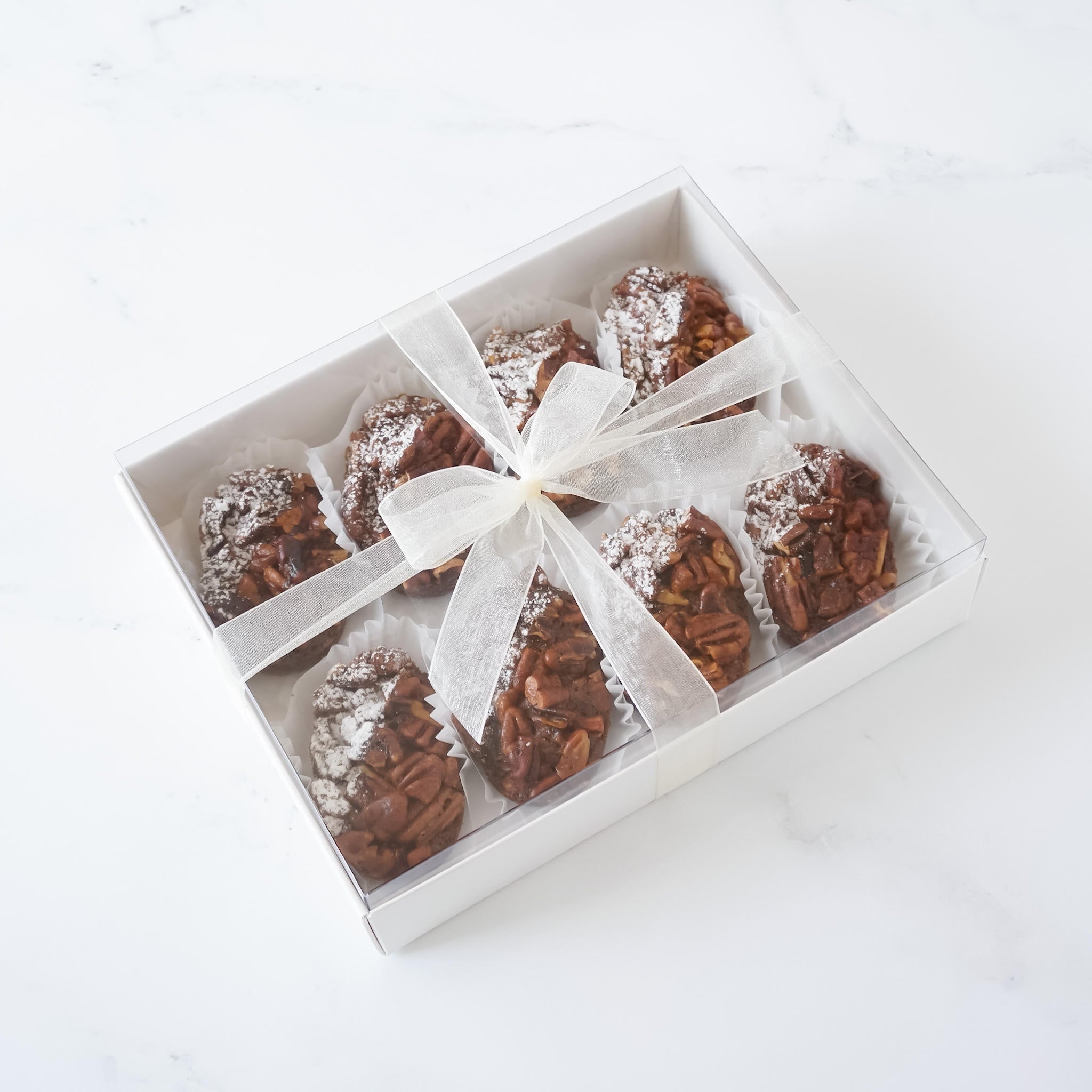 A white box with a clear lid containing eight chocolate pecan treats. The treats are arranged in two rows of four. The box is tied with a white ribbon and the treats are dusted with powdered sugar.