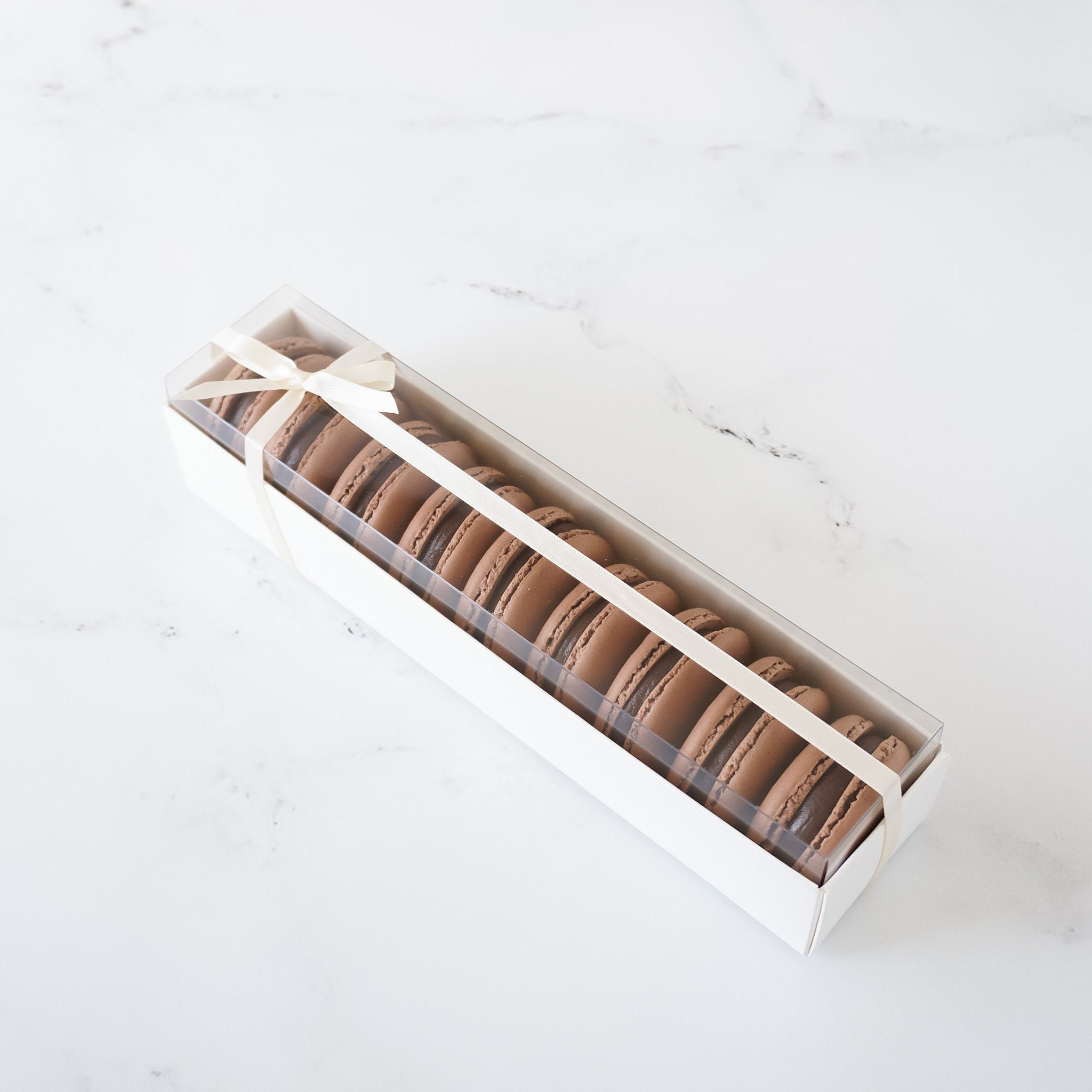 A white box with a white ribbon tied around it. The box is filled with chocolate macarons. The macarons are arranged in a row and are visible through the clear plastic lid of the box.