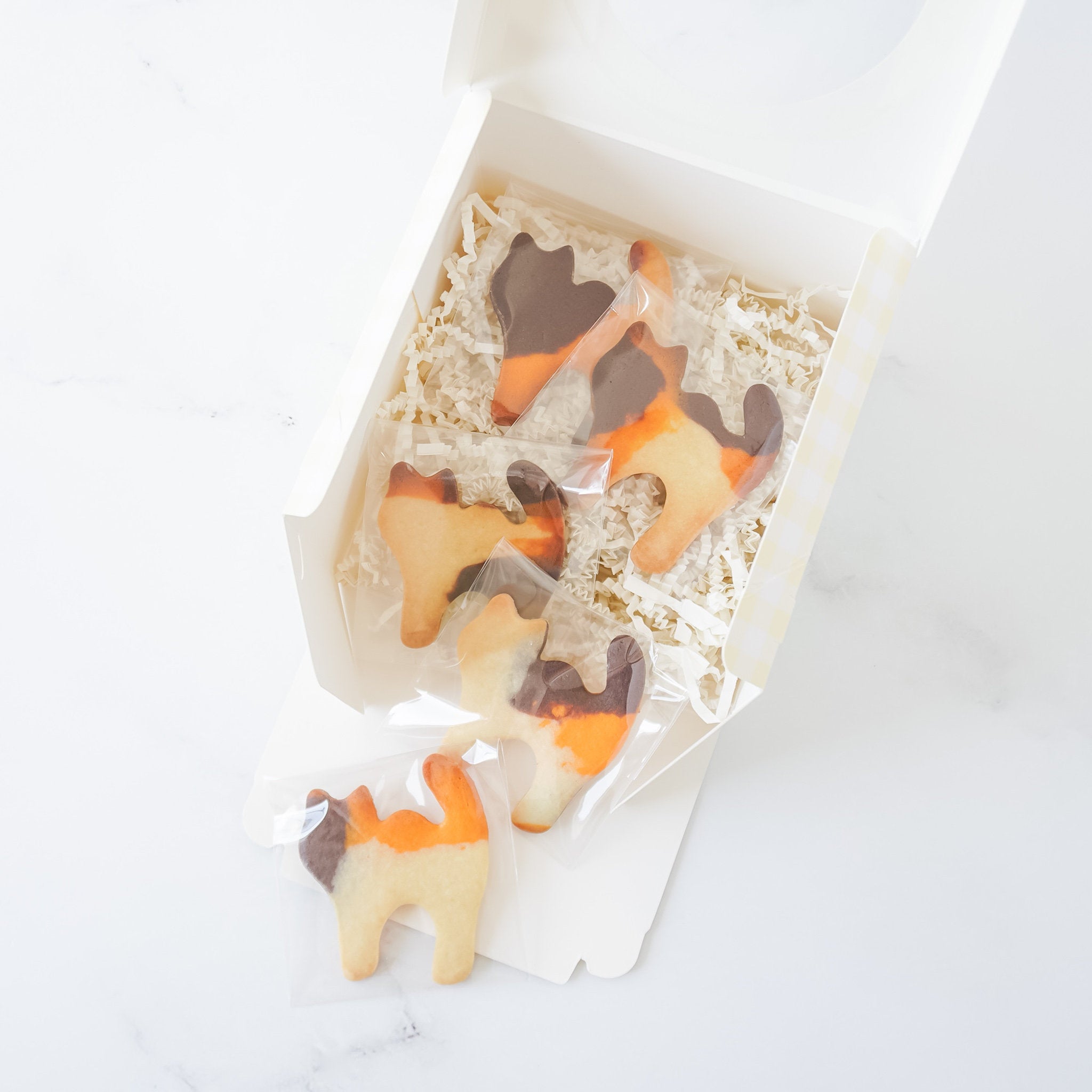 Five cat-shaped cookies in a white box with yellow gingham patterns. The cookies are decorated with orange, brown, and white icing. The cookies are in a white box with yellow gingham patterns. The box is open and the cookies are visible. The cookies are in a clear plastic bag.