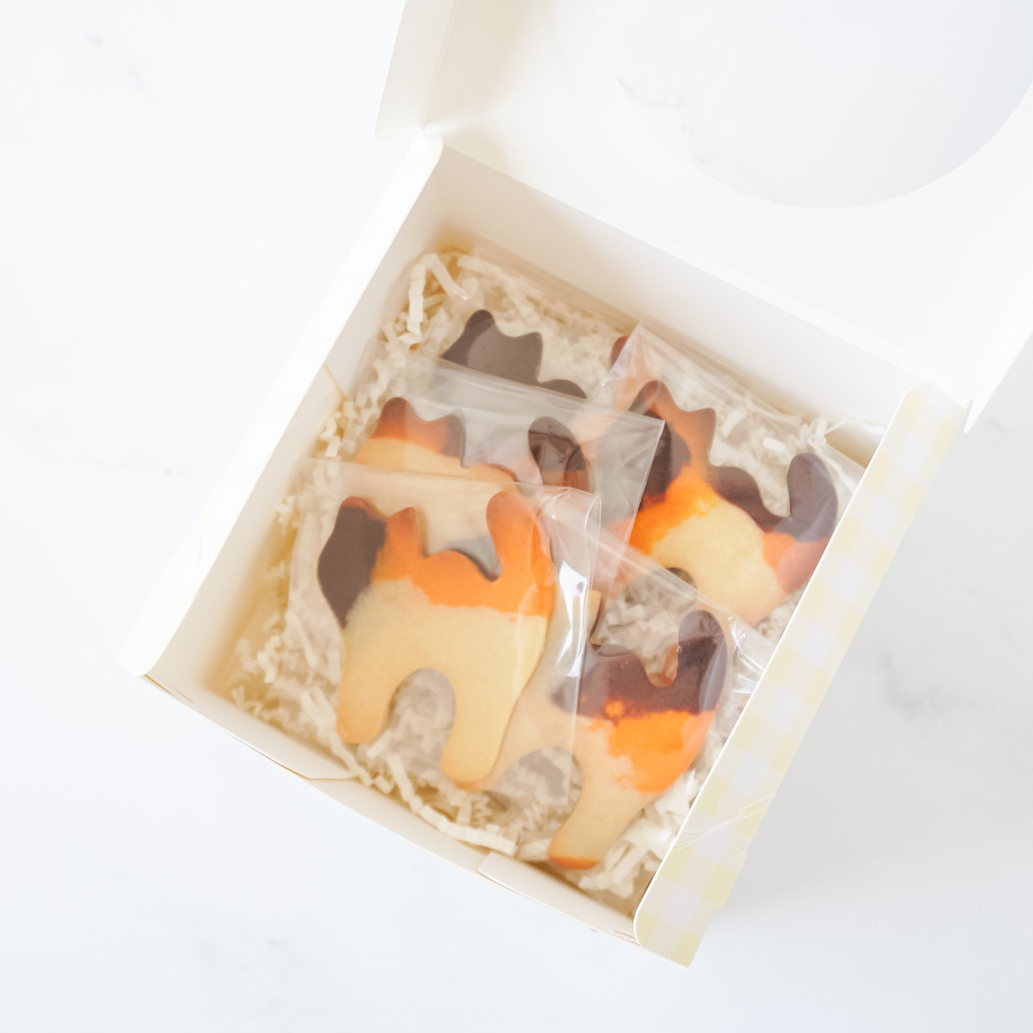 A white box with a clear window filled with five orange, brown, and white cat-shaped cookies. The cookies are individually wrapped in clear plastic bags.