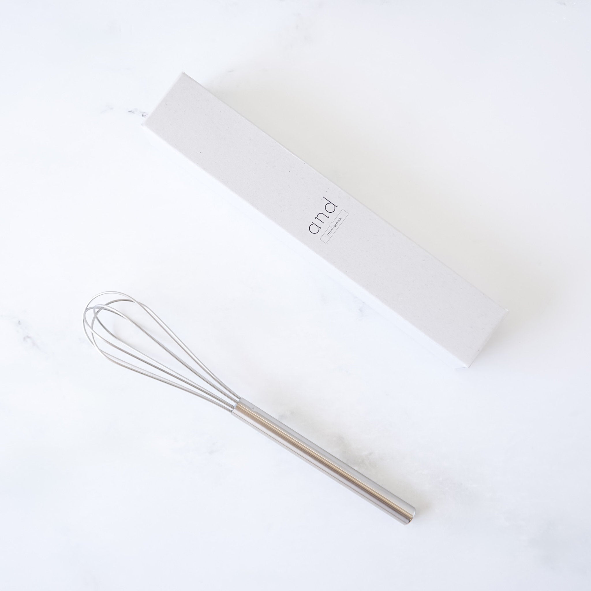 stainless steel mini whisk on a white surface with a branded box in the background