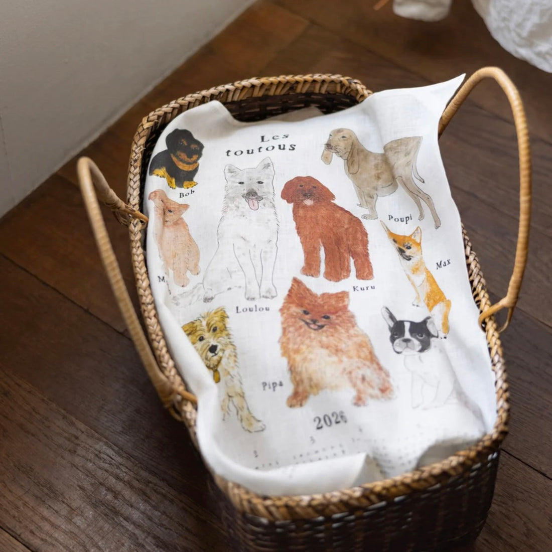 Wicker basket with a white kitchen cloth featuring illustrations of dogs on a wooden floor.