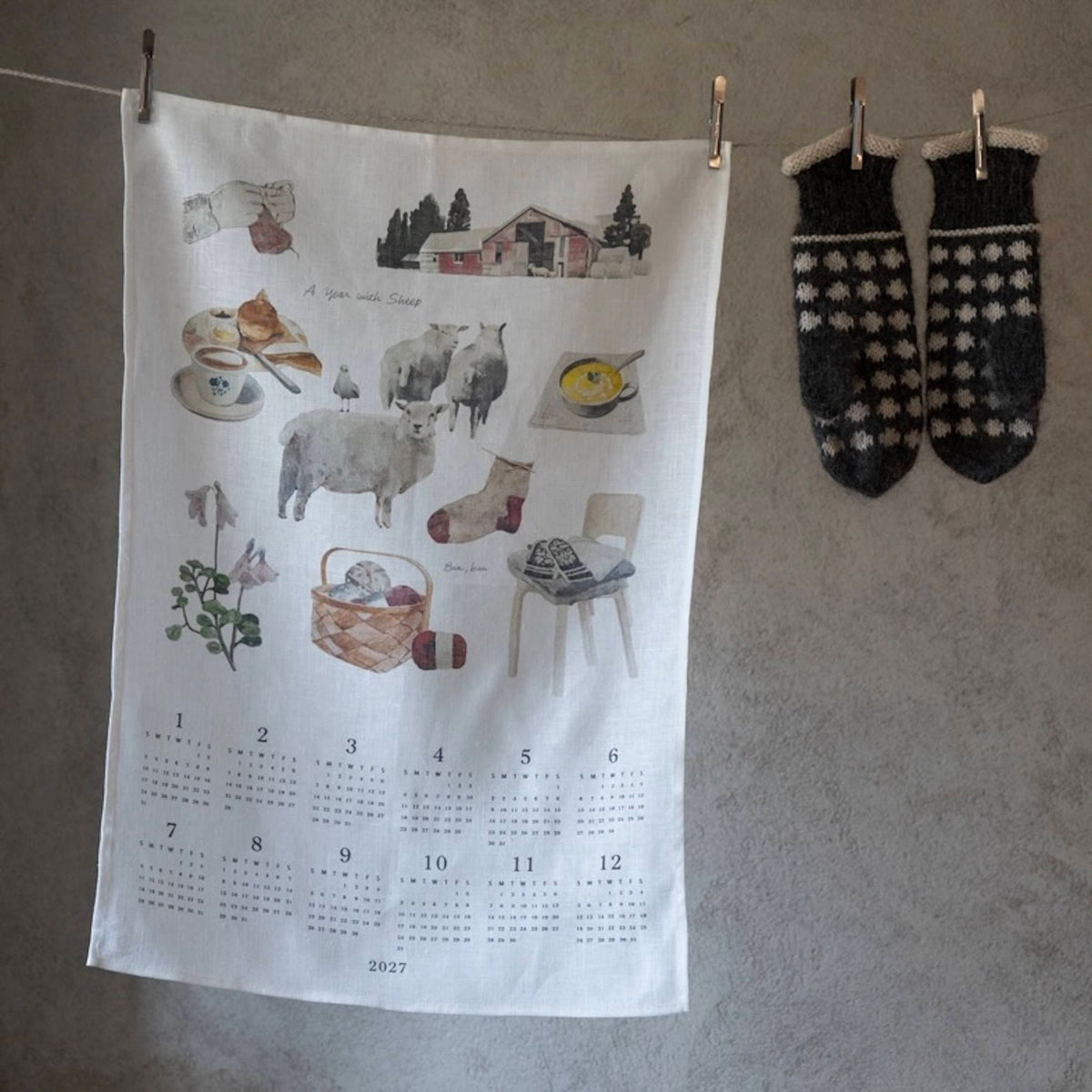 A white fabric wall calendar towel hanging on a gray wall, clipped to a string. The towel features a watercolor-style illustration of cozy winter scenes, including sheep, a red barn, a mug, a basket, and a chair, with a 12-month grid at the bottom labeled 2027.
