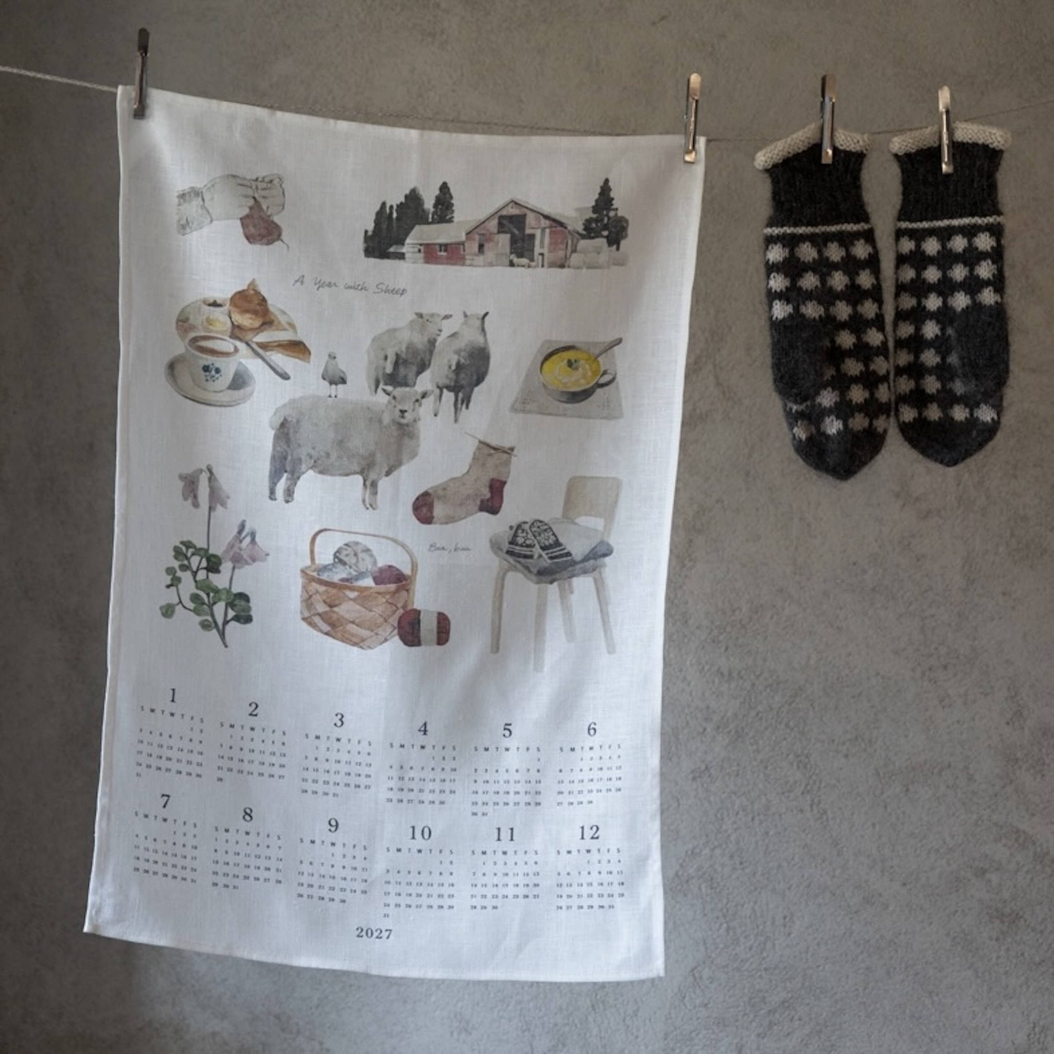 A white fabric wall calendar towel hanging on a gray wall, clipped to a string. The towel features a watercolor-style illustration of cozy winter scenes, including sheep, a red barn, a mug, a basket, and a chair, with a 12-month grid at the bottom labeled 2027.