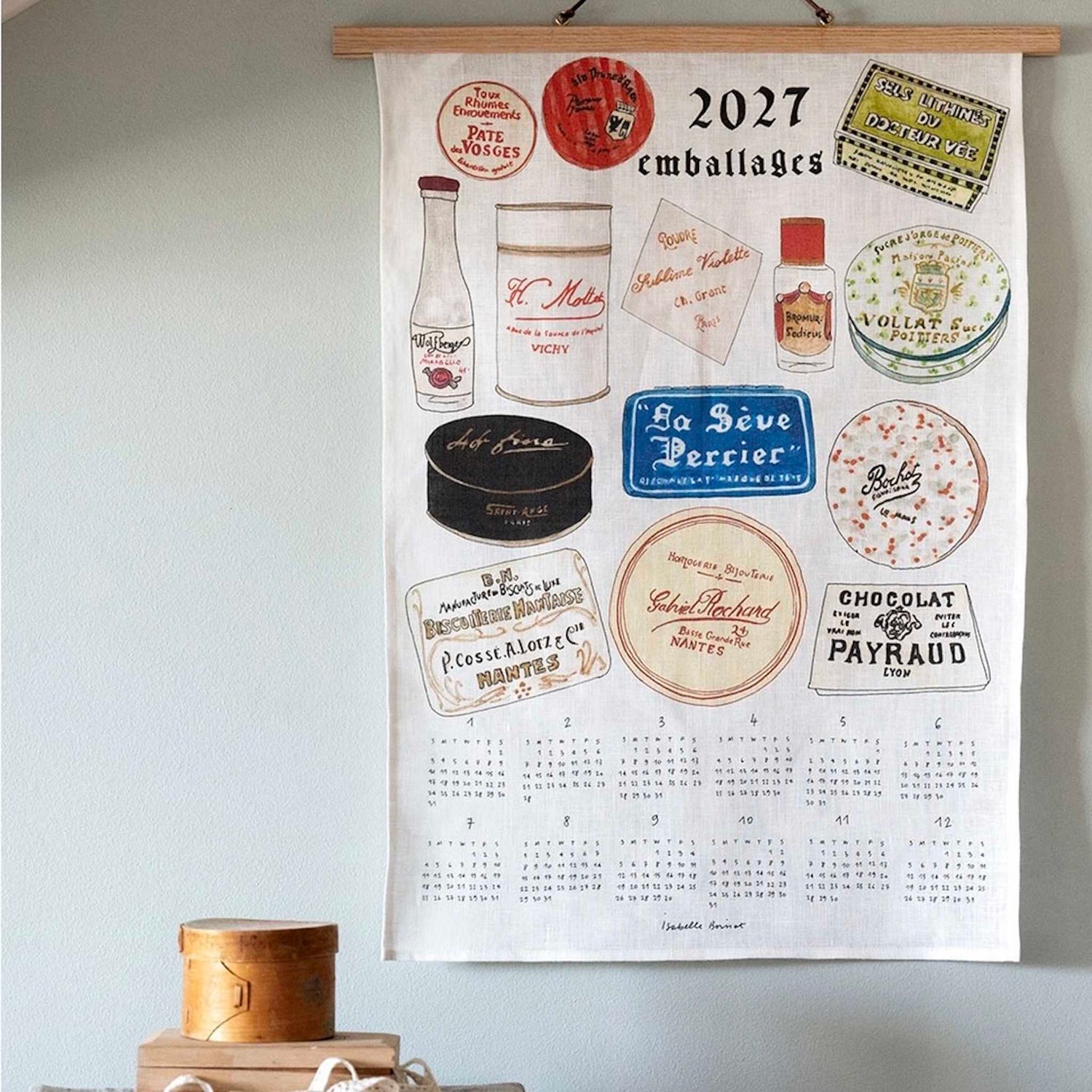 A wooden-framed wall calendar hanging on a light blue wall, featuring vintage French candy and cosmetic packaging illustrations with the text &
